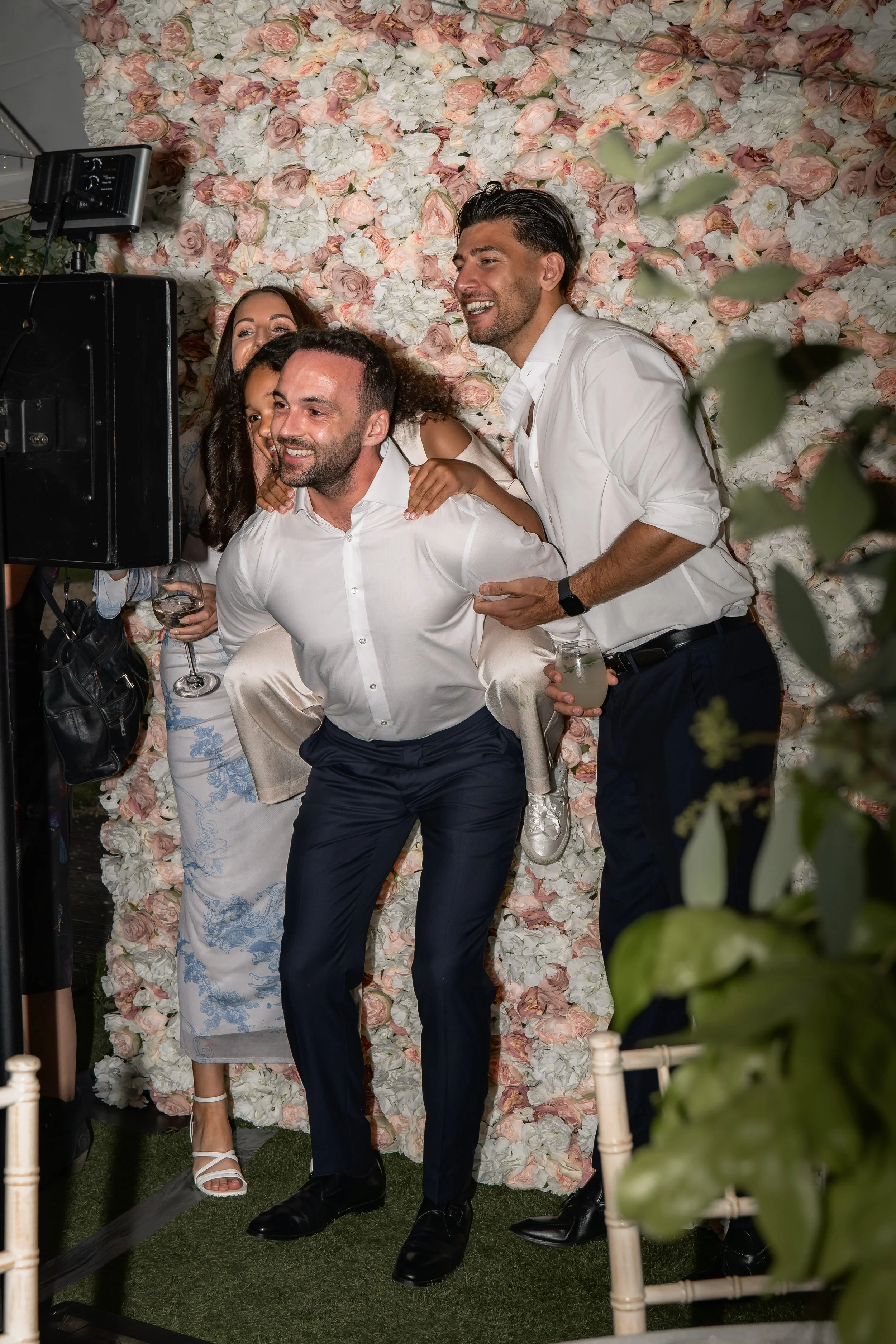 Group of friends at a celebration, posing for a photo in front of a floral backdrop, with some holding drinks.