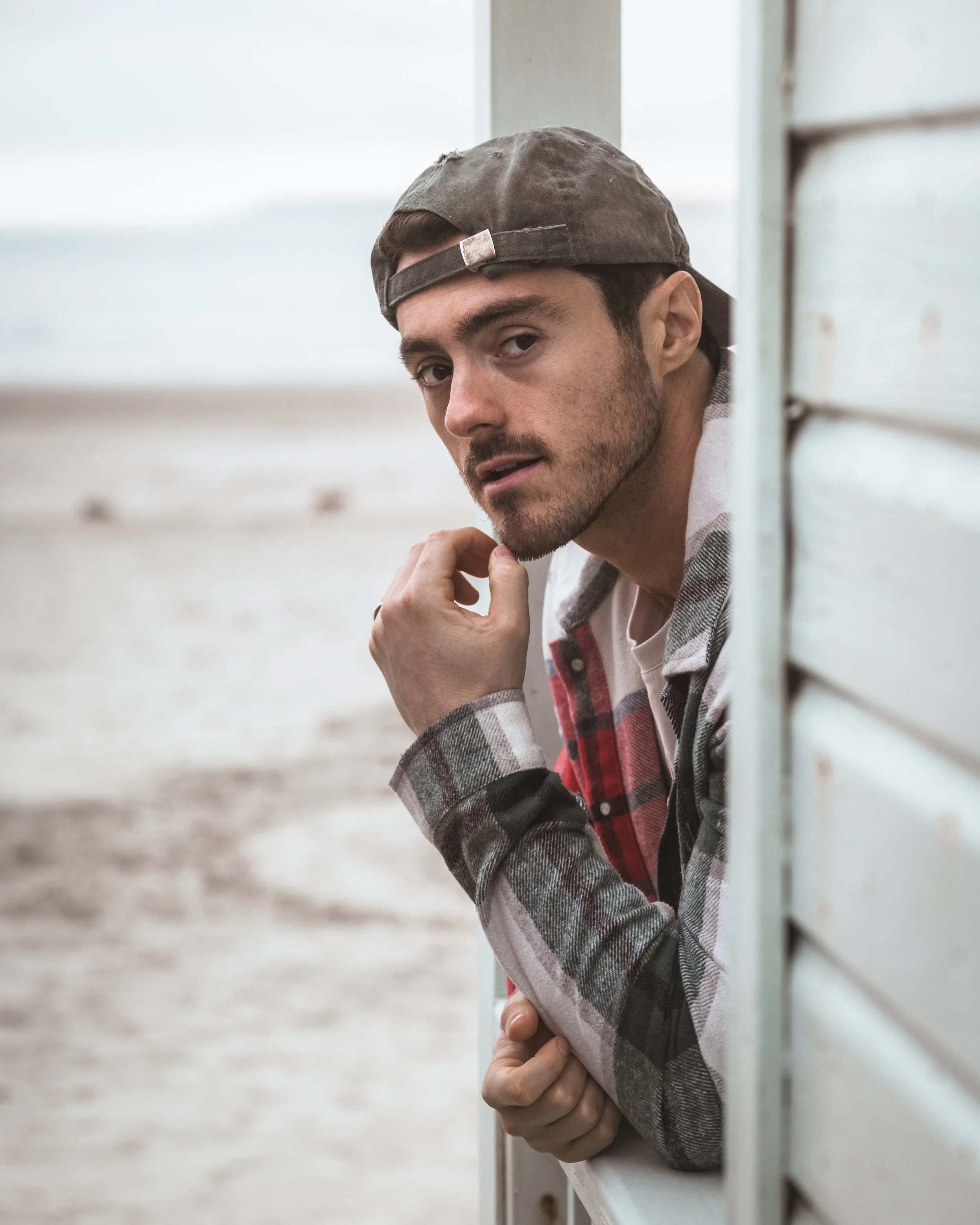 Man wearing a baseball cap and plaid shirt leaning out of a small window, looking at the camera.