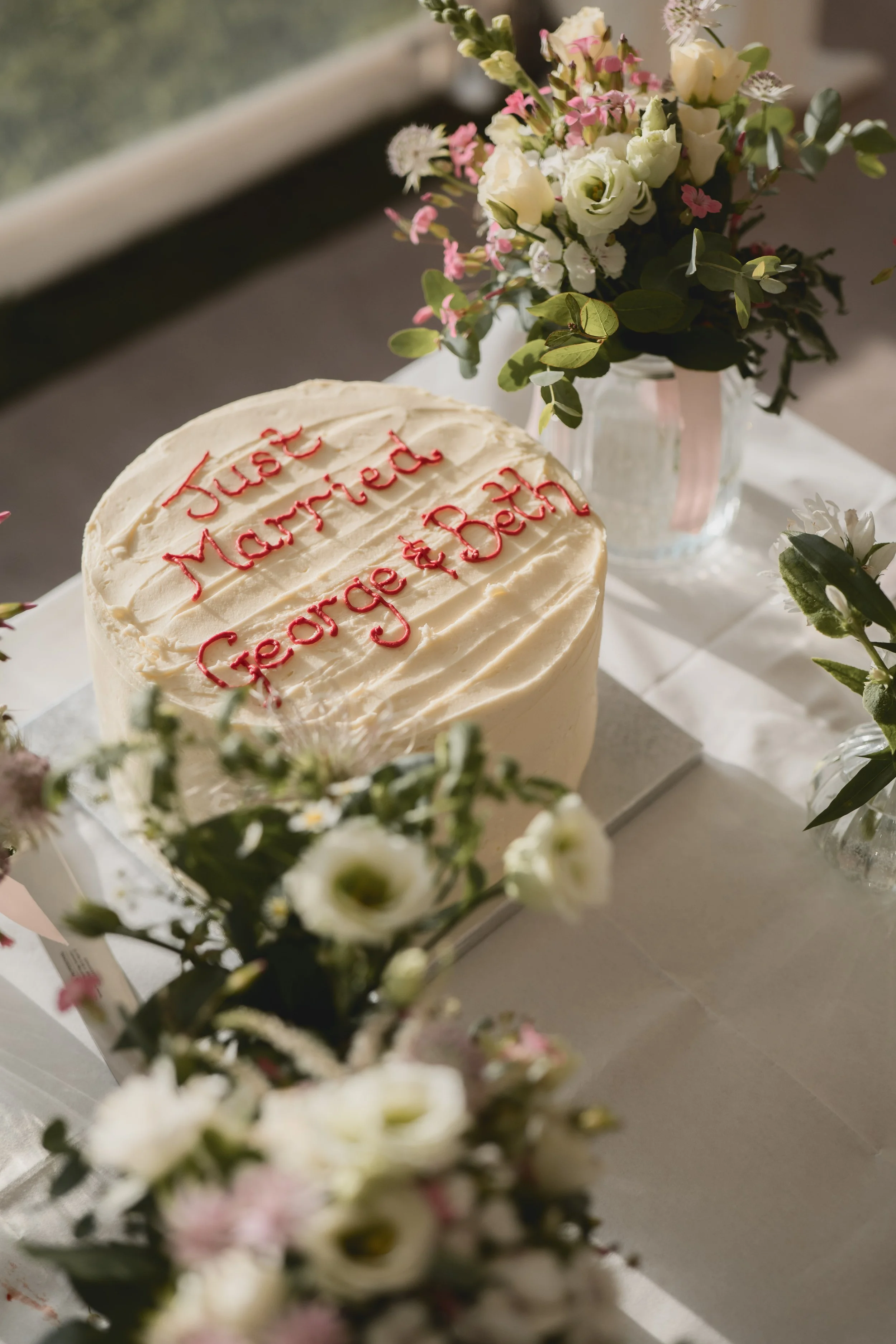 A wedding cake with white frosting and red writing reading 'Just Married George & Beth', surrounded by floral arrangements in vases on a table.