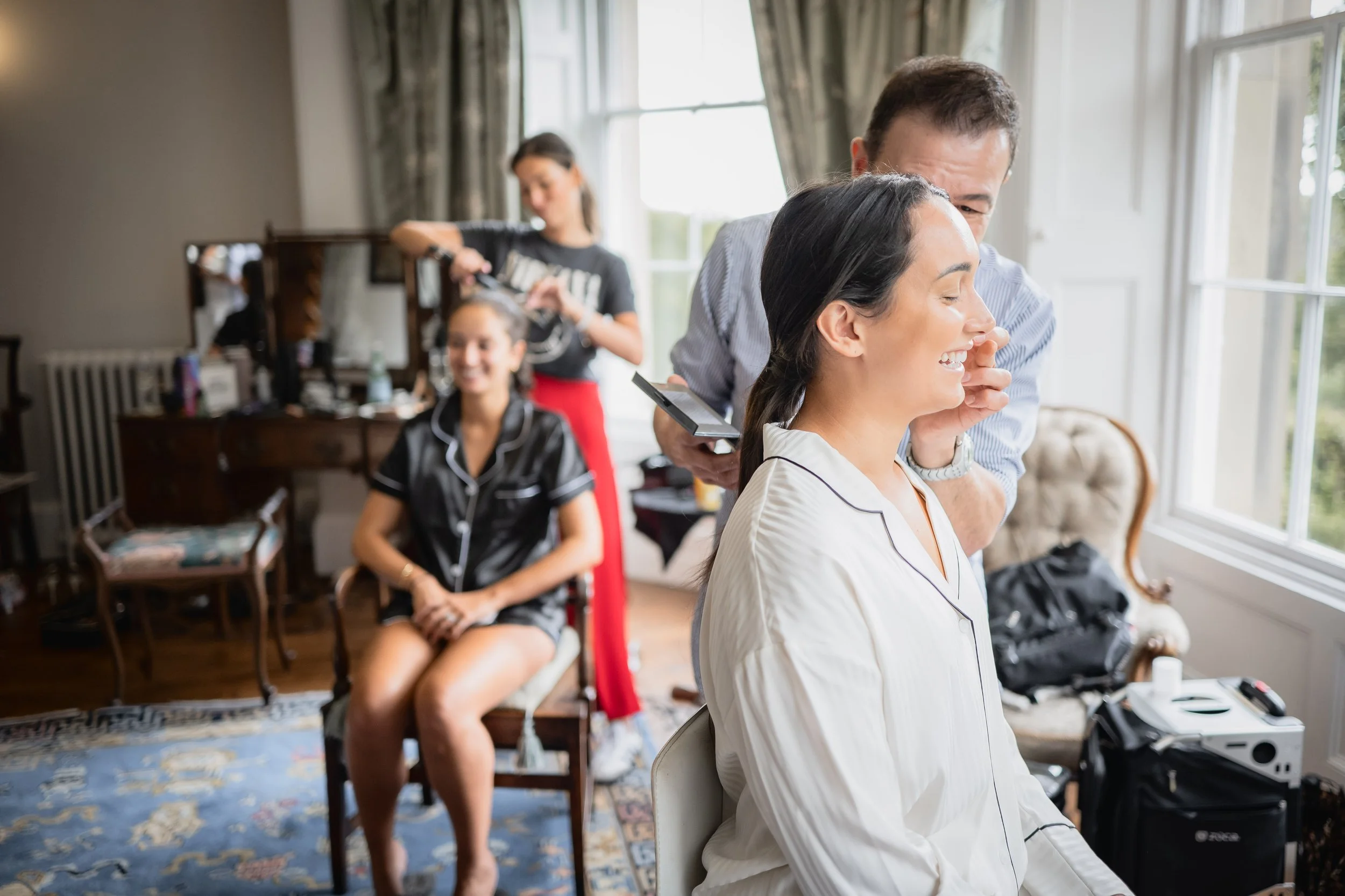 A woman in pajamas is having her hair styled by a man while smiling. In the background, a makeup artist is working on another woman seated in a chair.