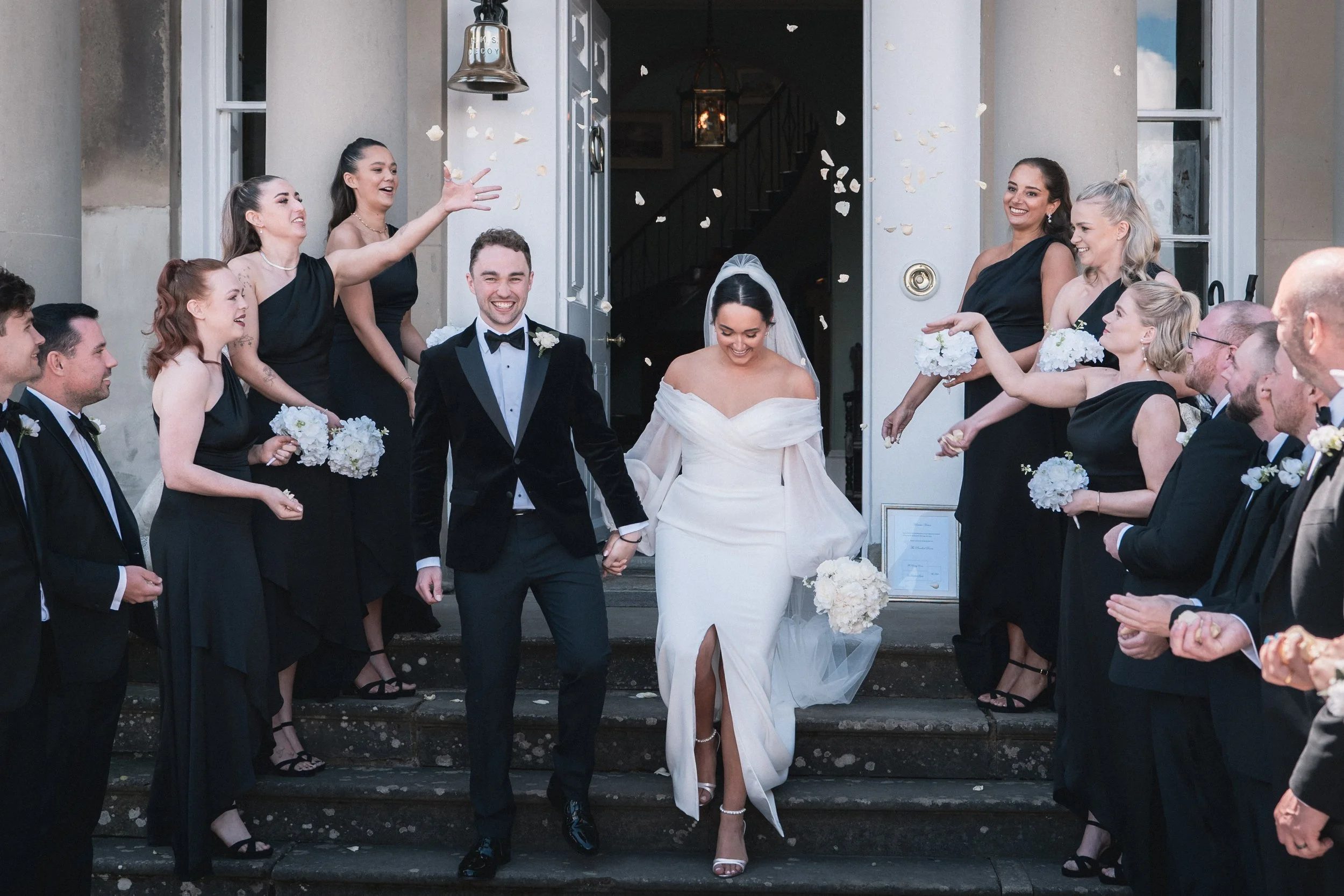 A bride and groom walking out of a building while wedding guests throw flower petals.