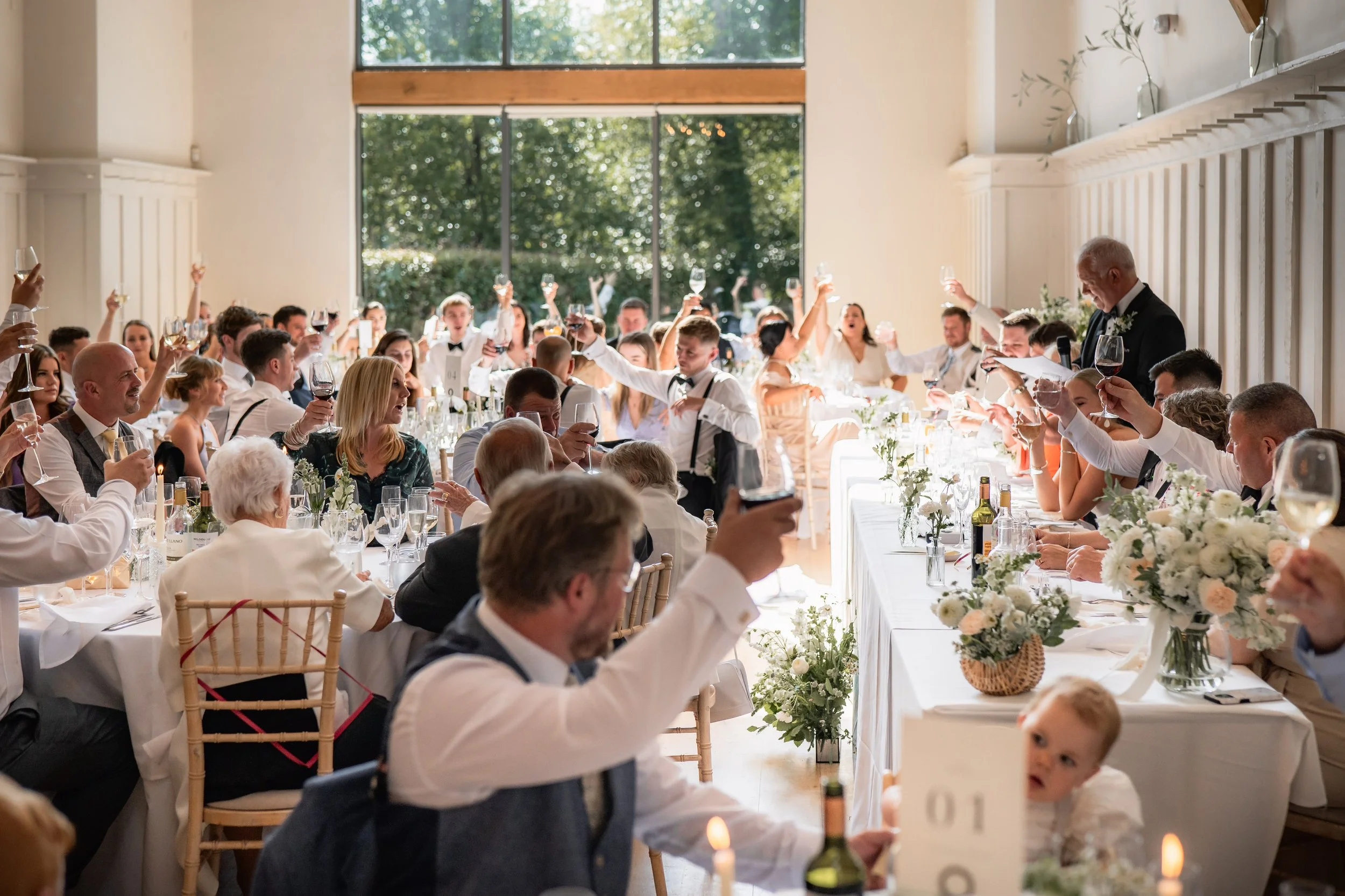 Guests at a wedding reception raising glasses in a toast inside a bright, airy venue with large windows and greenery outside.