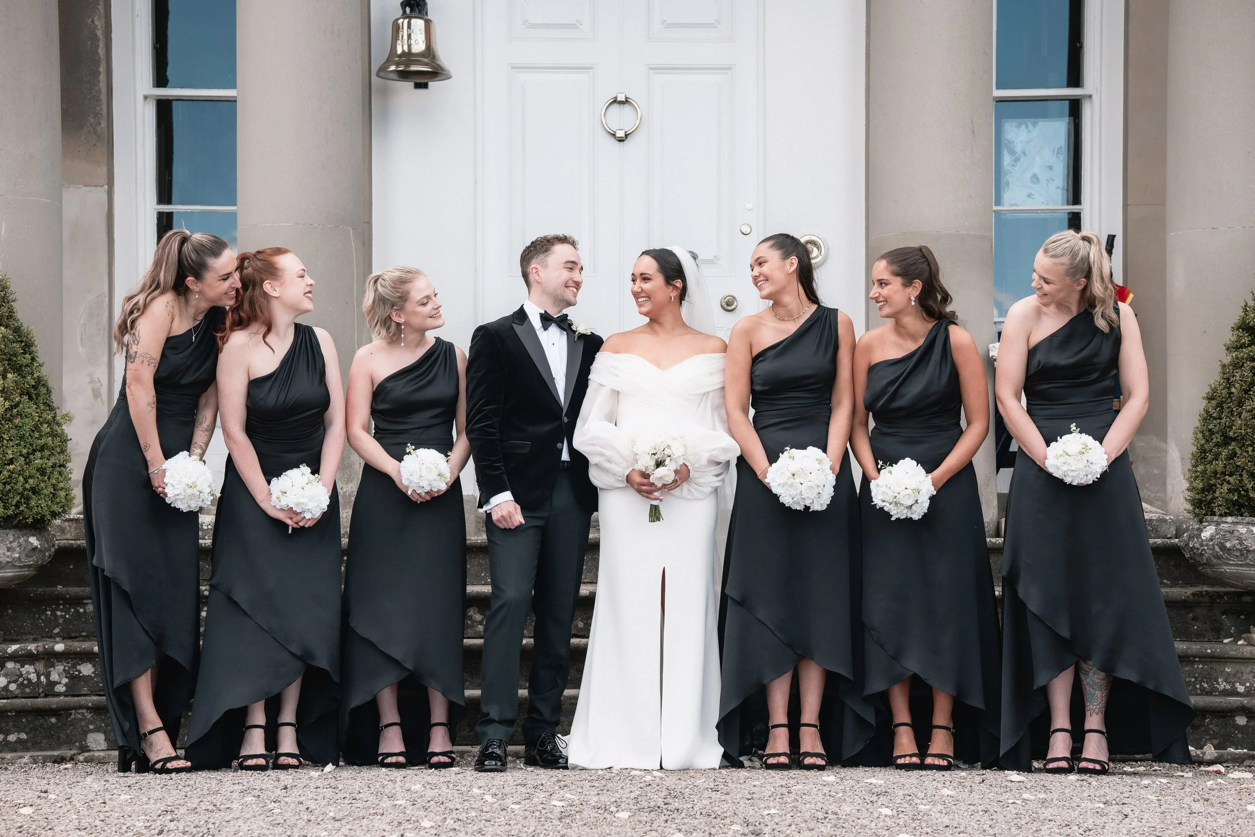 A wedding party standing on steps outside a building, with the bride and groom in the center. The bride is wearing a white dress holding a bouquet, and the groom is in a black tuxedo. Six bridesmaids in black dresses holding white bouquets are standi