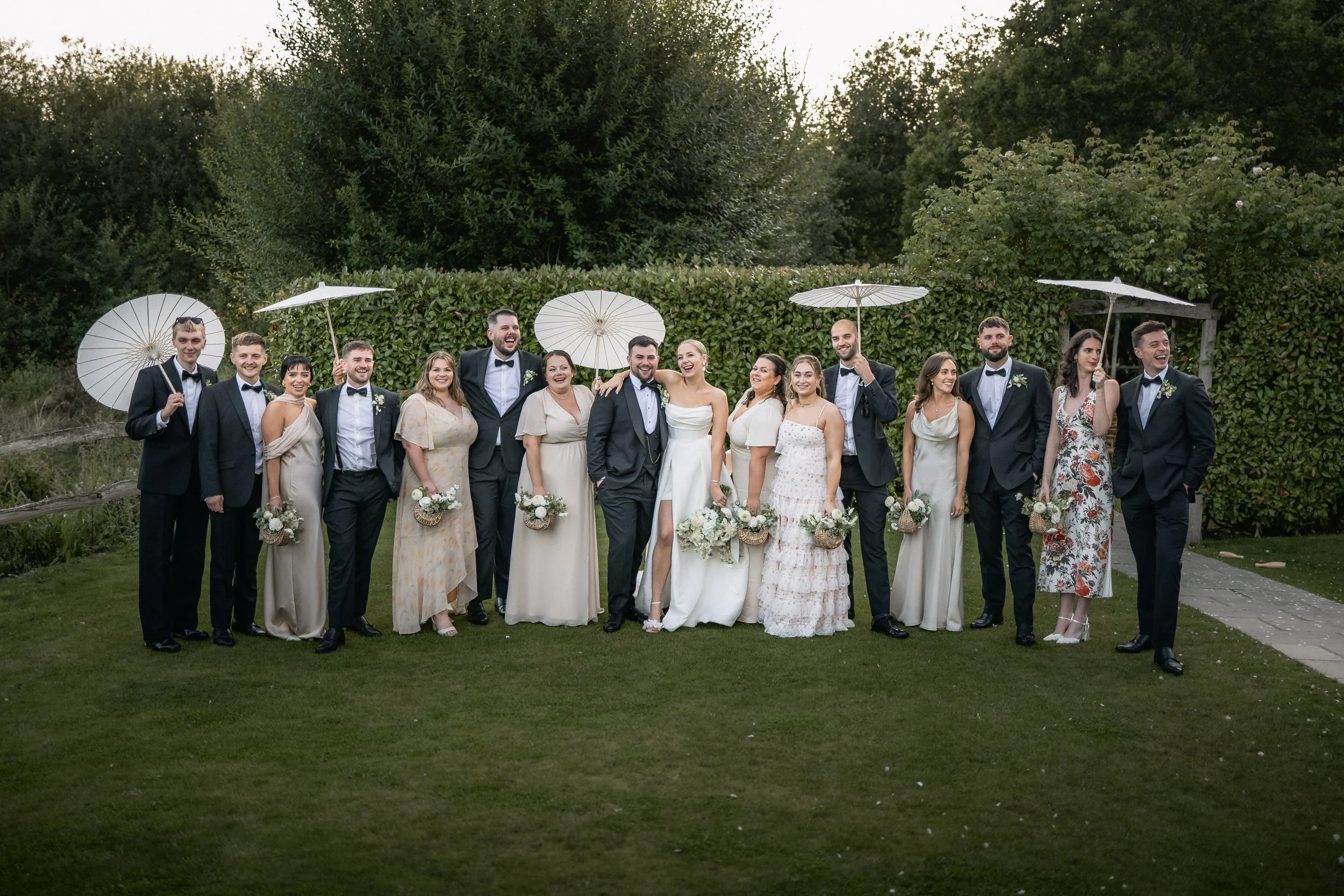 Group of wedding guests, including the bride and groom, standing outdoors on a grassy area with trees and bushes in the background. Some guests are holding white parasols, and several are holding floral bouquets. The group is dressed in formal attire