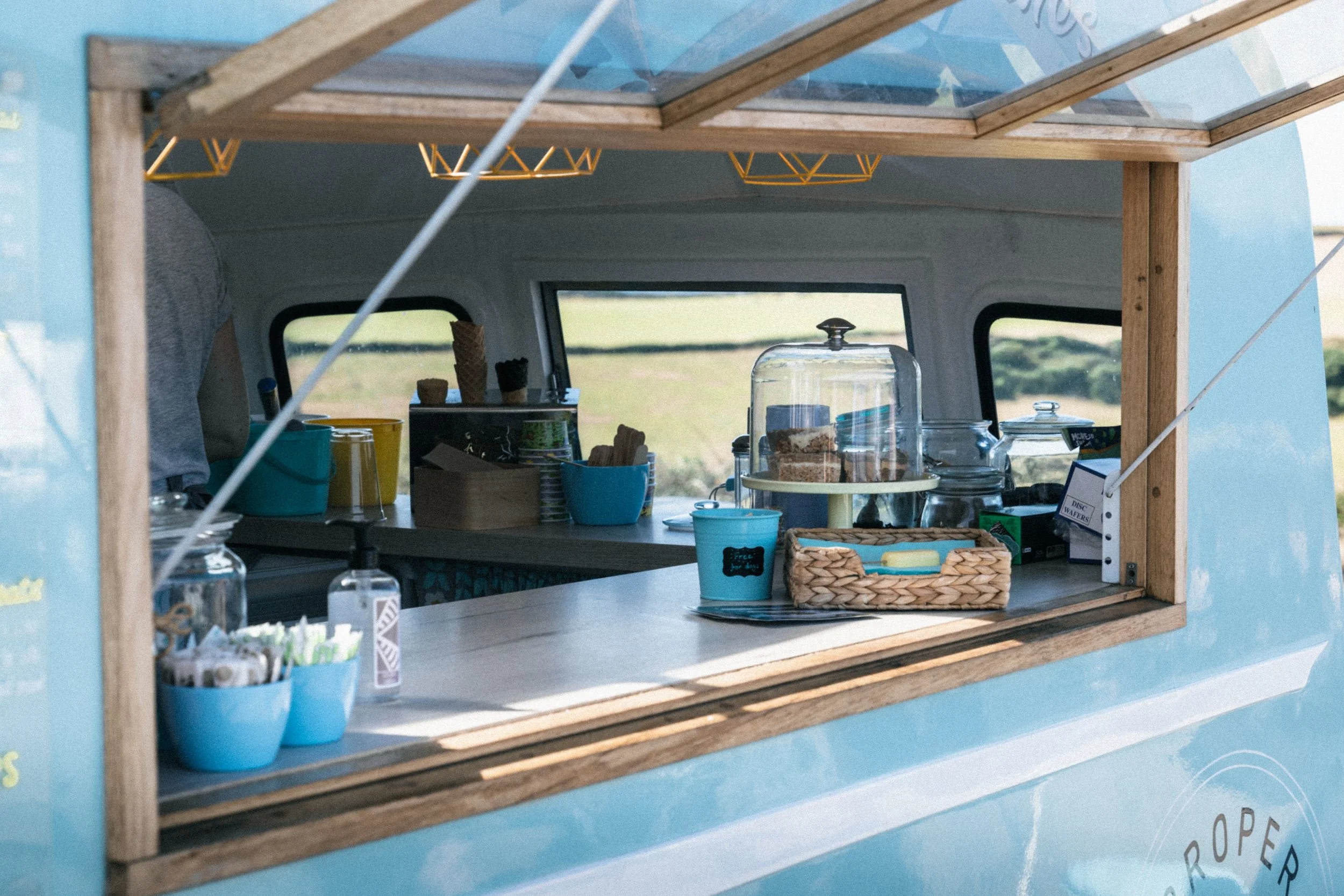 Inside a light blue food truck with a wooden window frame, showing a counter with cookies, cake slices under a glass dome, and coffee cups, with windows revealing a grassy field outside.