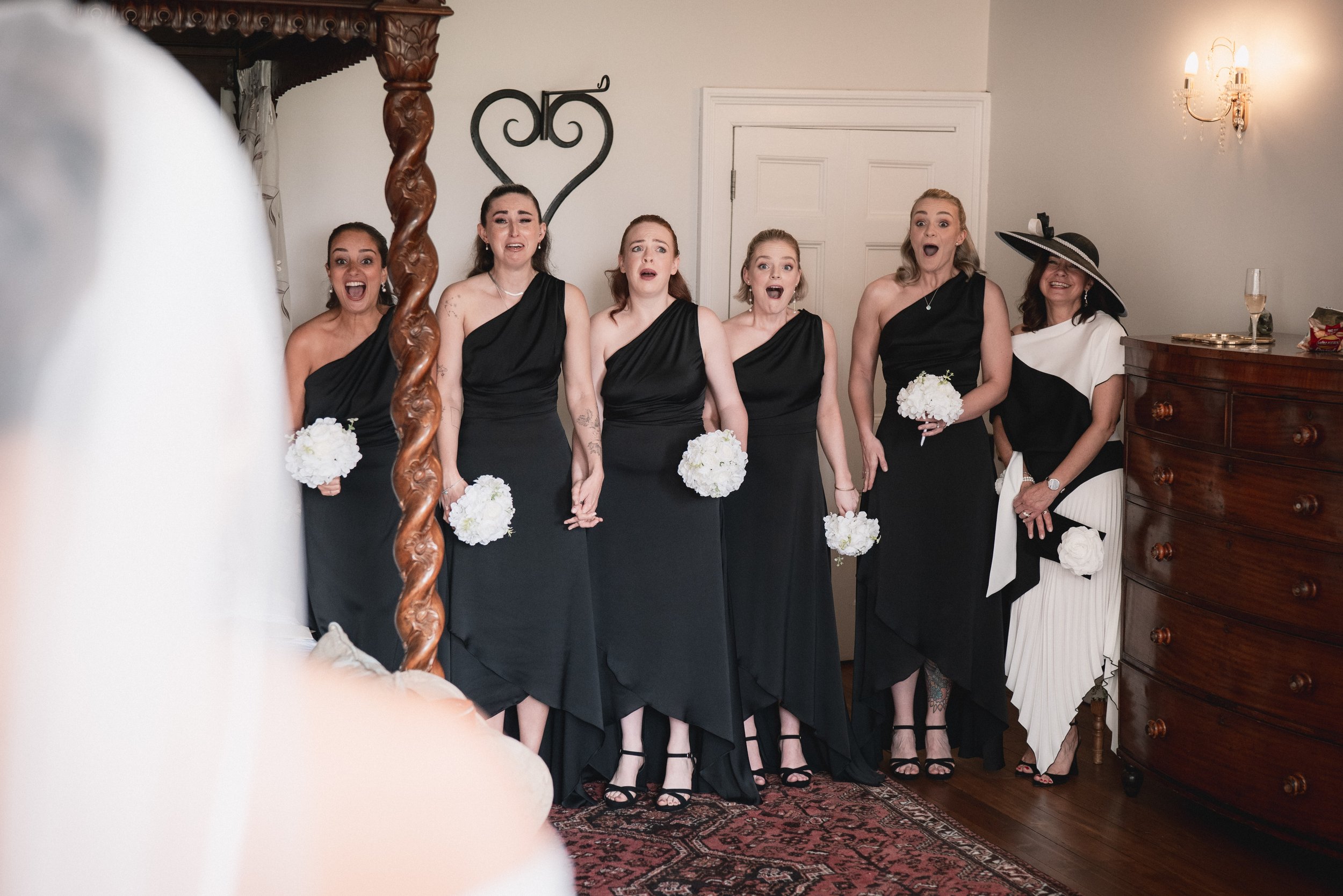 Group of six women dressed in elegant black and white dresses, holding white flower bouquets, reacting with surprise or shock, indoors with a wooden dresser and chandelier visible.