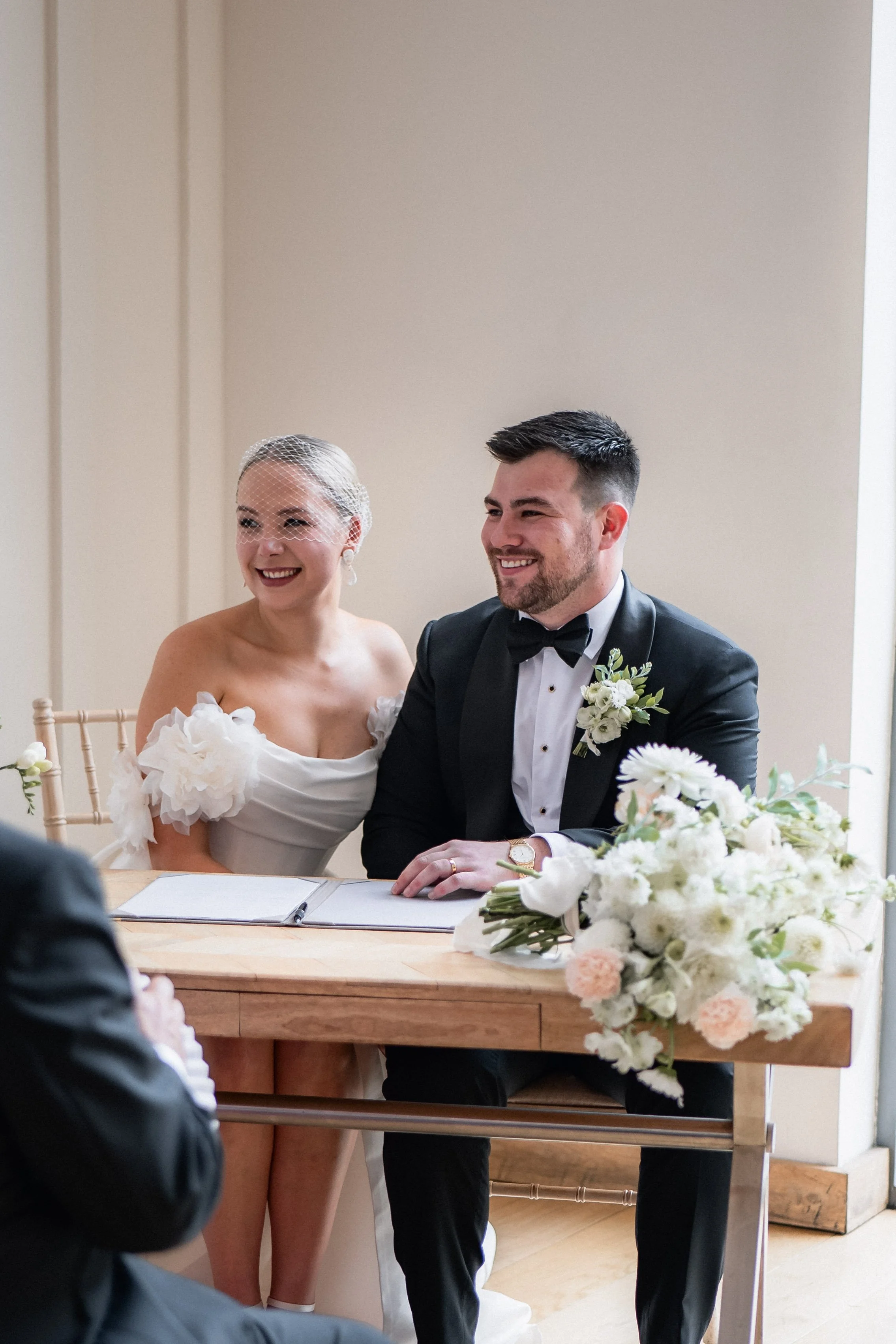 A bride and groom sitting at a table during their wedding ceremony, smiling and holding hands, with a bouquet of white and pink flowers on the table.
