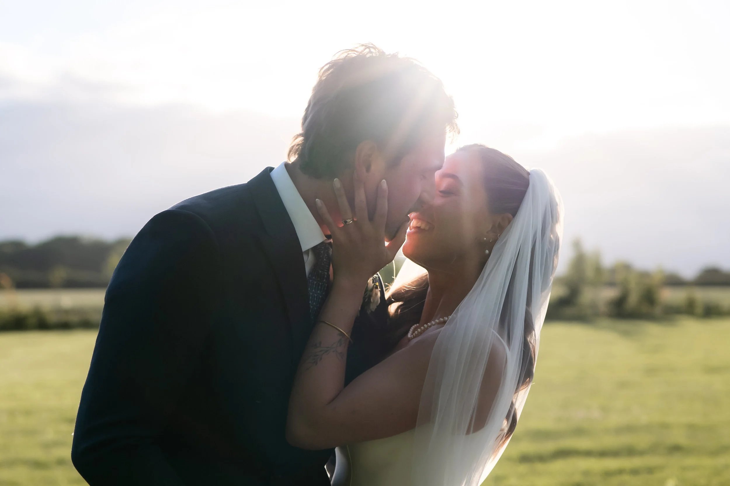 A newlywed couple sharing a kiss outdoors during sunset, with the groom in a black suit and the bride in a white dress and veil, overlooking a grassy field.