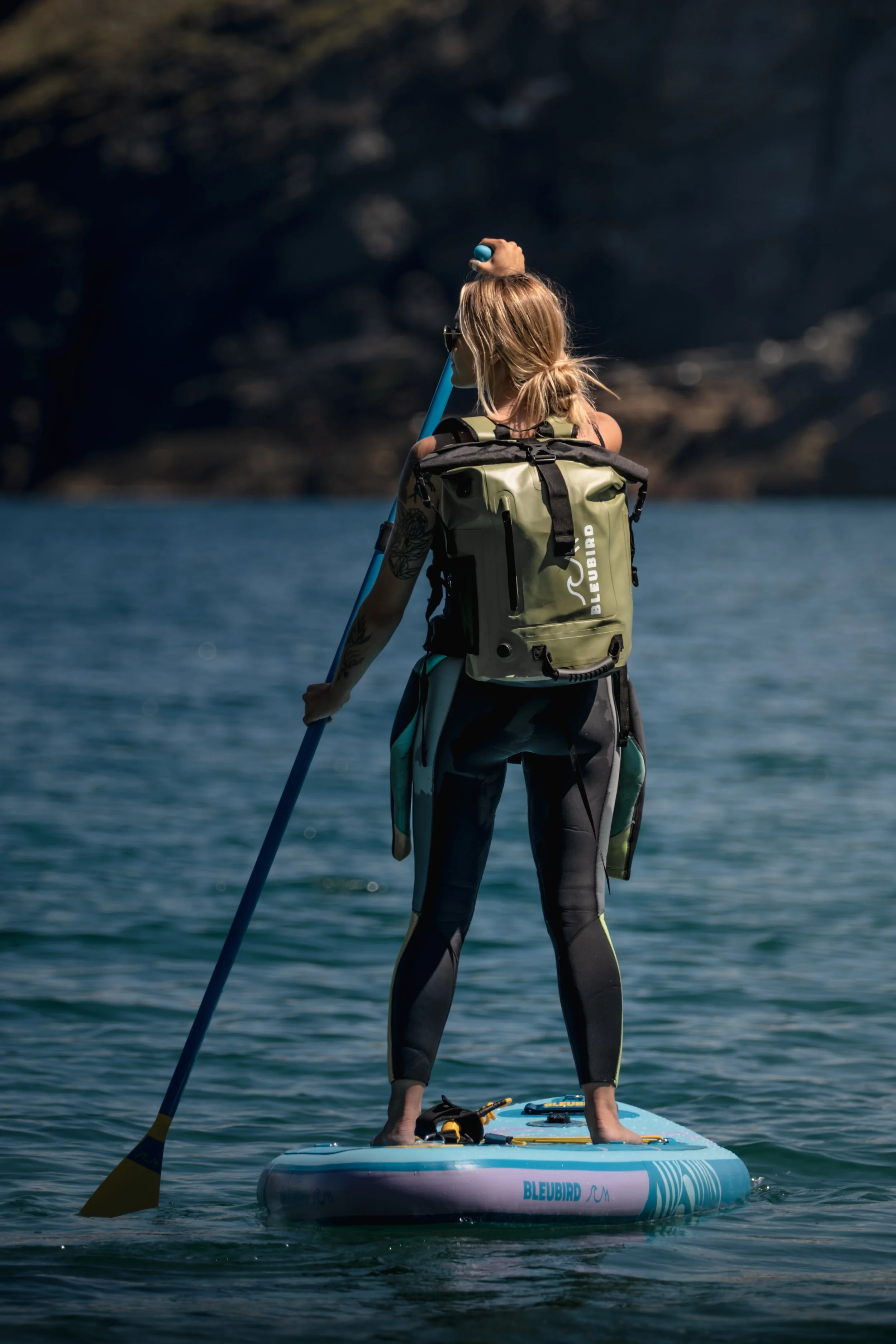 A woman standing on a stand-up paddleboard on a body of water, holding a paddle and wearing a backpack and wetsuit.