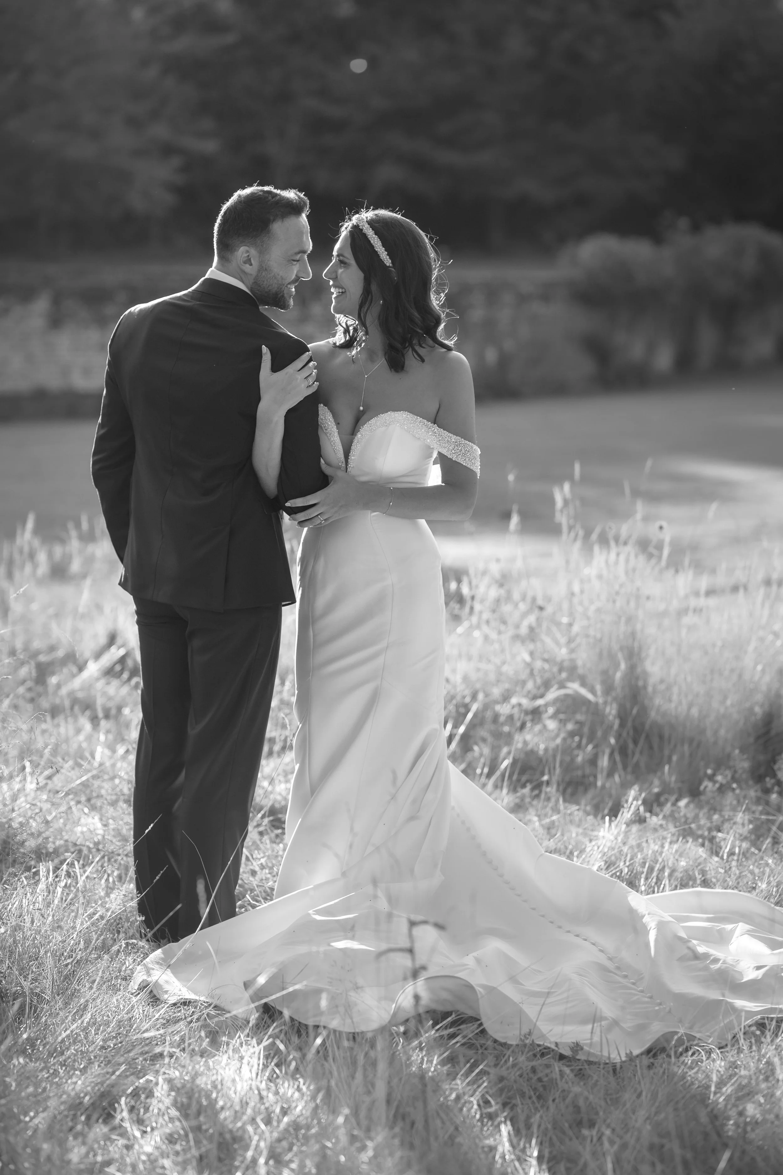 A black and white photo of a bride and groom standing outdoors on a grassy field, smiling and gazing at each other, with a lake and trees in the background.