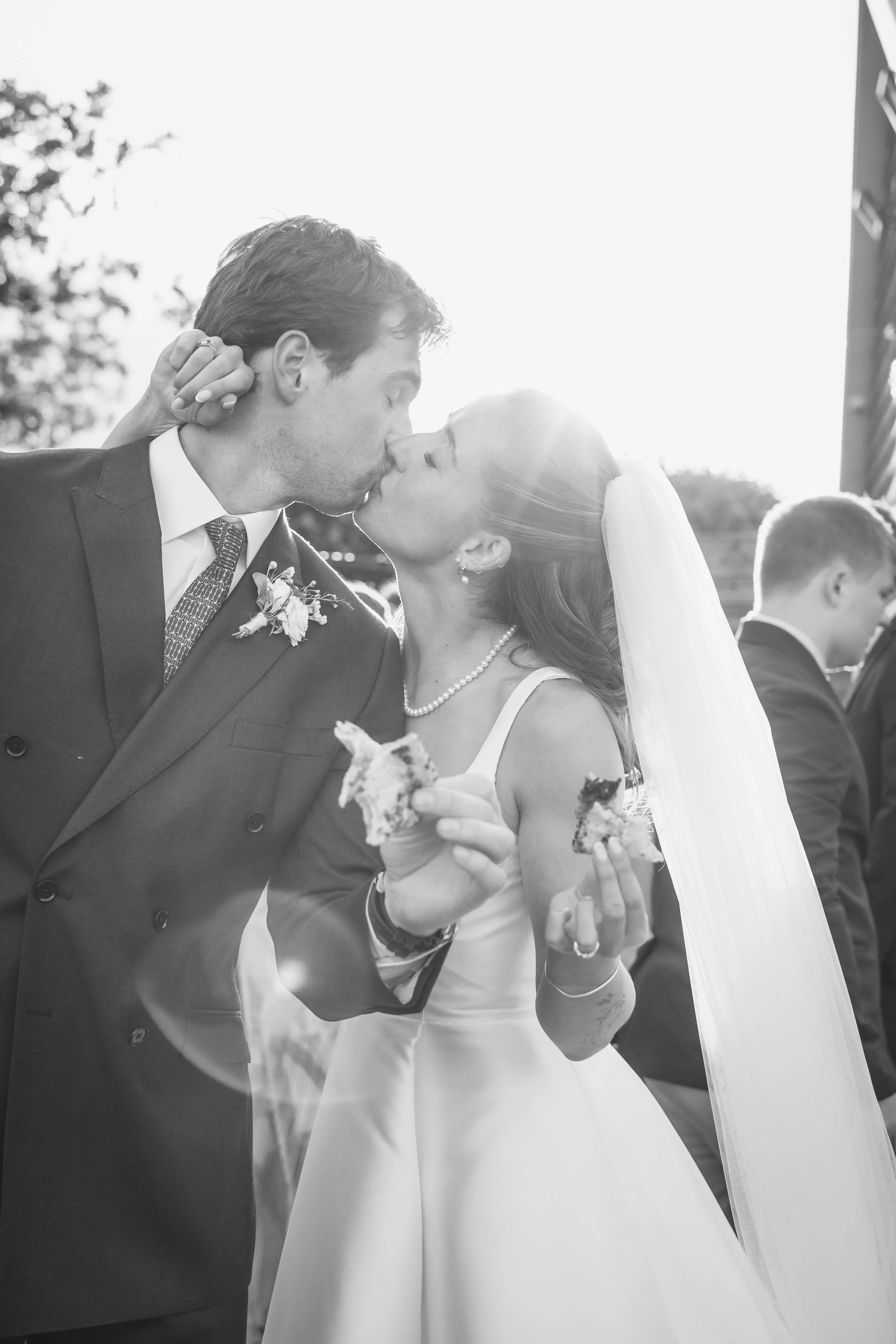 A black and white photo of a newlywed couple kissing outdoors, the man in a suit and the woman in a wedding dress with a veil, holding pieces of cake.