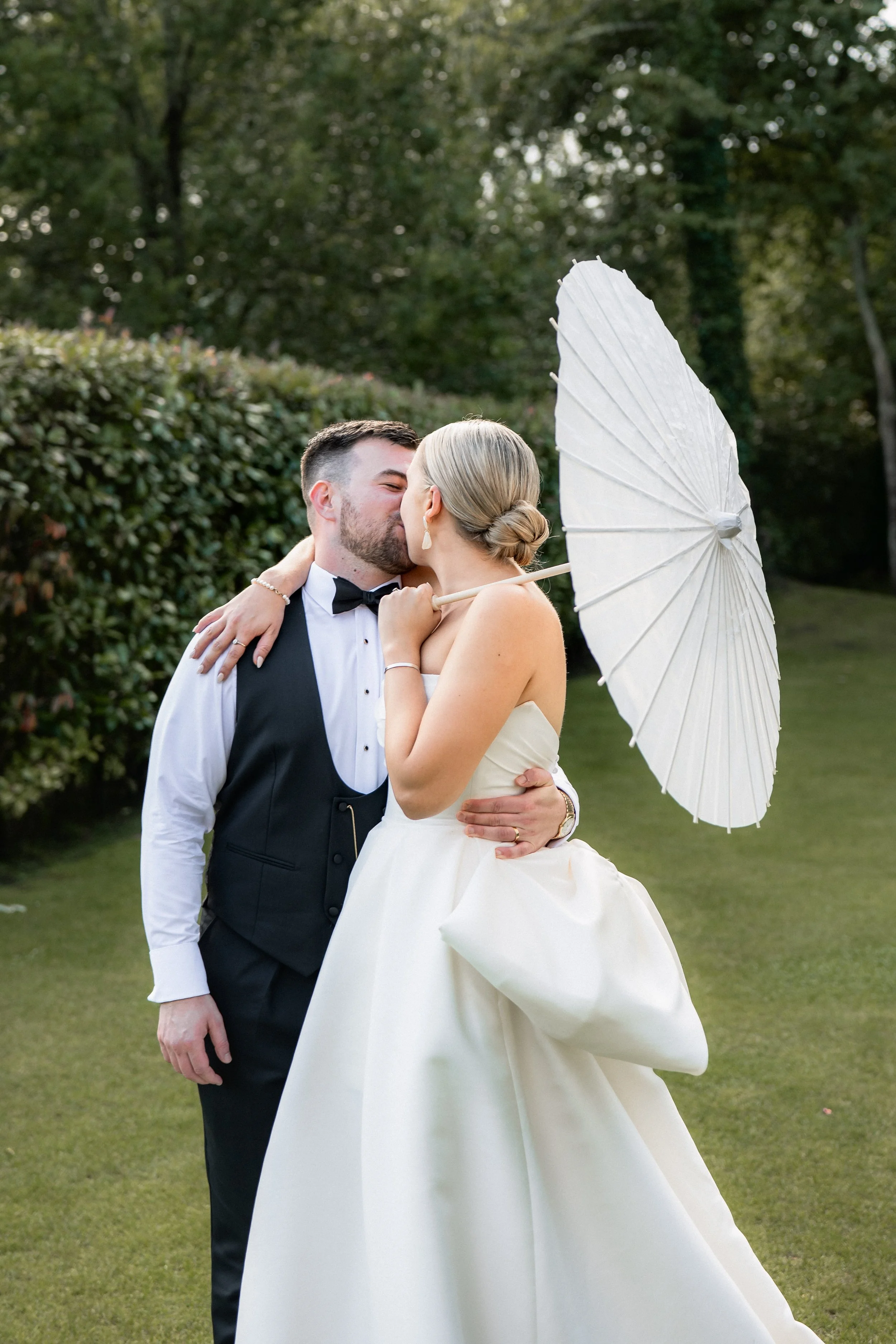 A newlywed couple sharing a kiss outdoors, the bride holding a white parasol, with trees and greenery in the background.