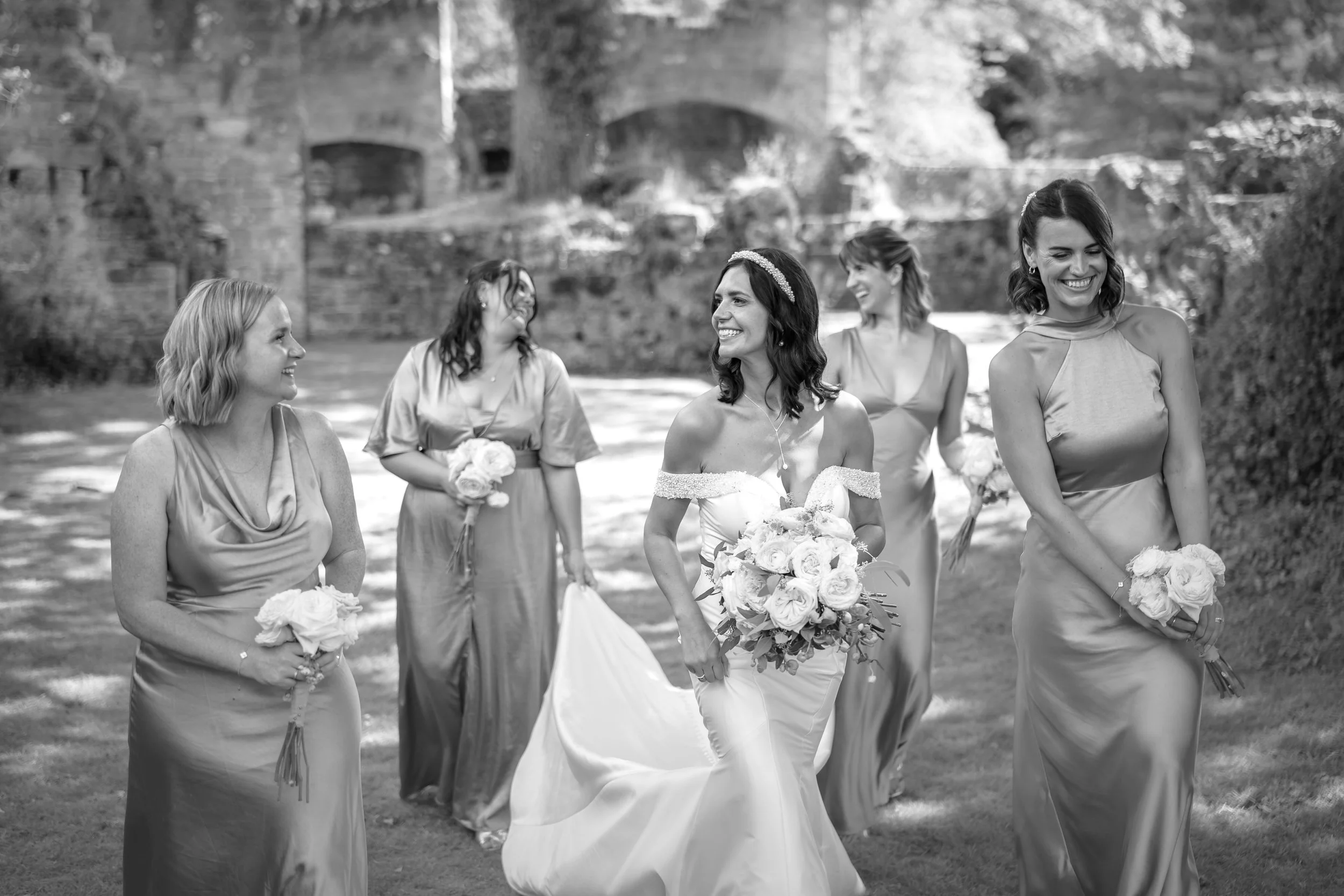 Black and white photo of a bride and her bridesmaids walking outdoors, smiling and holding bouquets. The bride is in the center, wearing a wedding dress, with four bridesmaids in gowns around her.