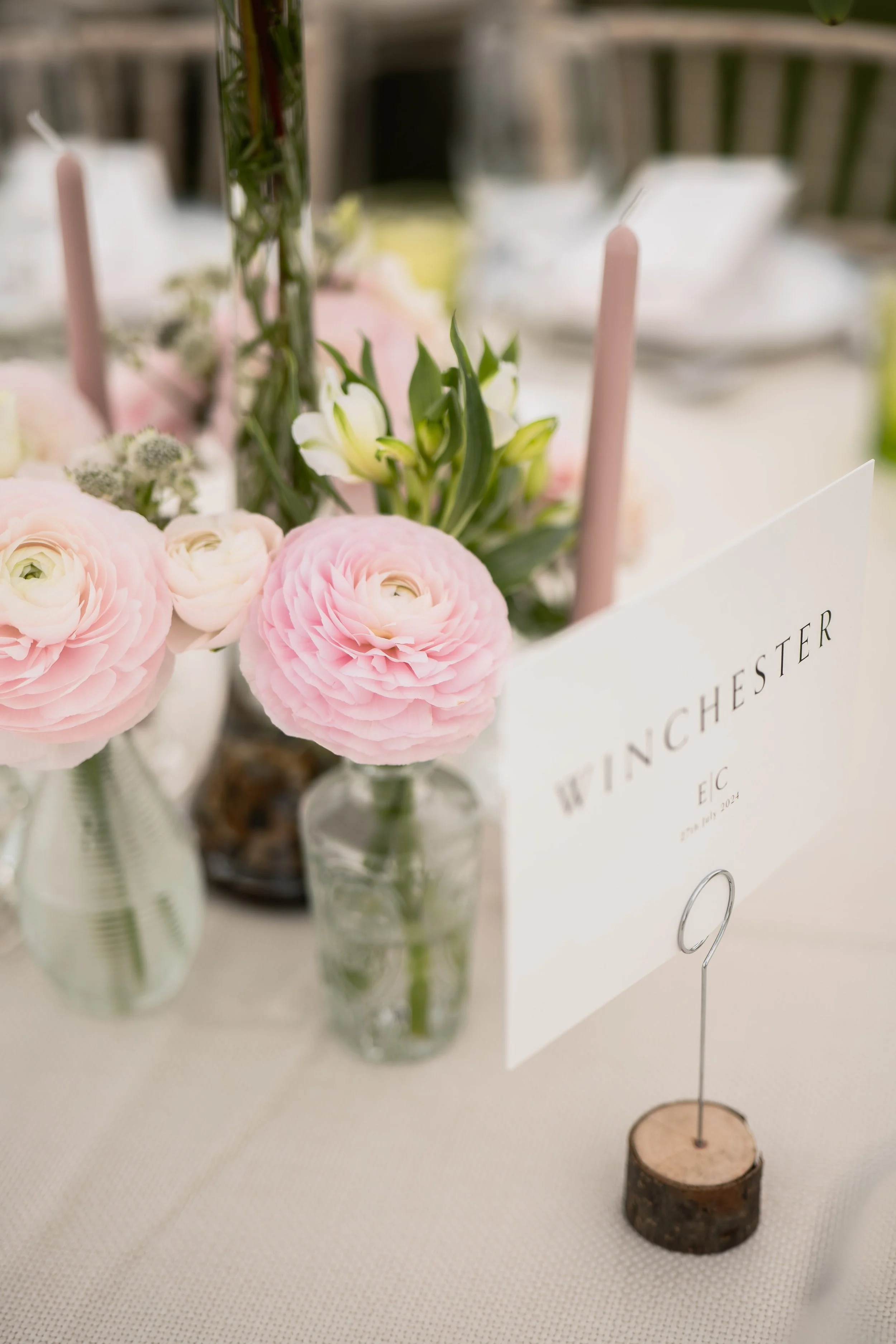 A close-up of a floral centerpiece with pink and white flowers, a table sign with the words "WINCHESTER," and pink taper candles on a white tablecloth at a wedding reception.