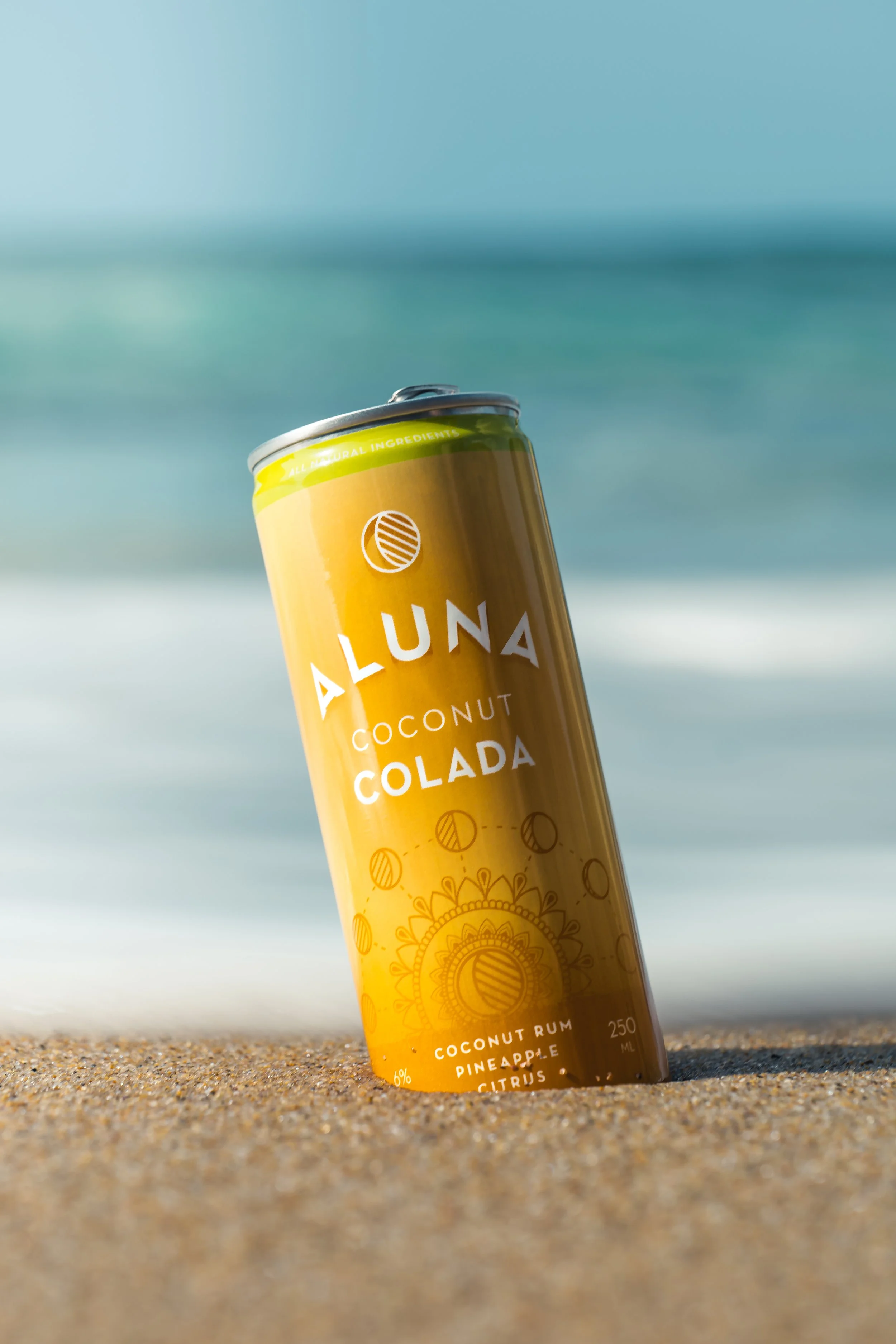 A can of Aluna Coconut Colada drink in the sand at the beach with ocean and sky in the background.
