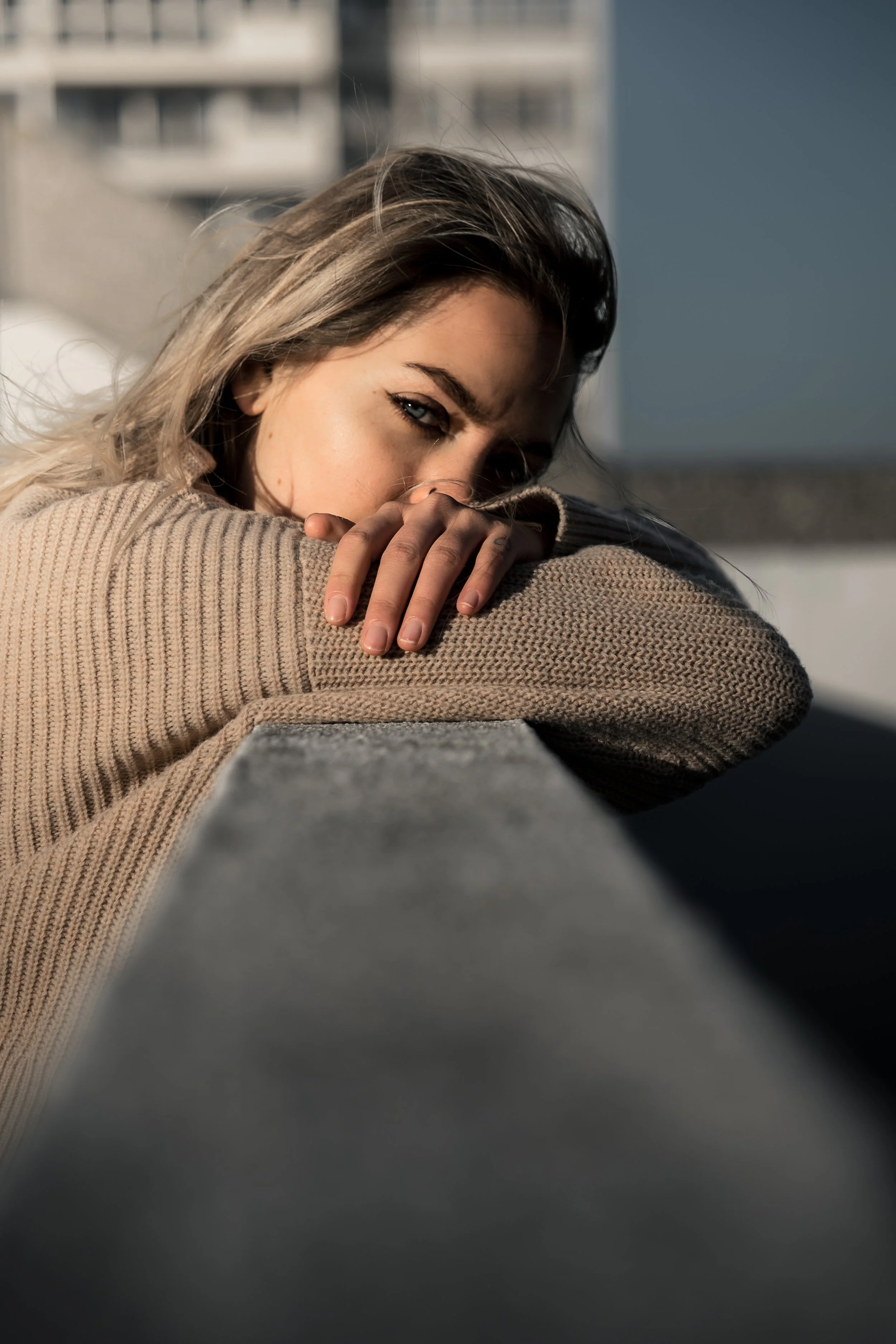 A young woman with blonde hair leaning on a concrete ledge, wearing a beige sweater, looking over her arm with an intense gaze.