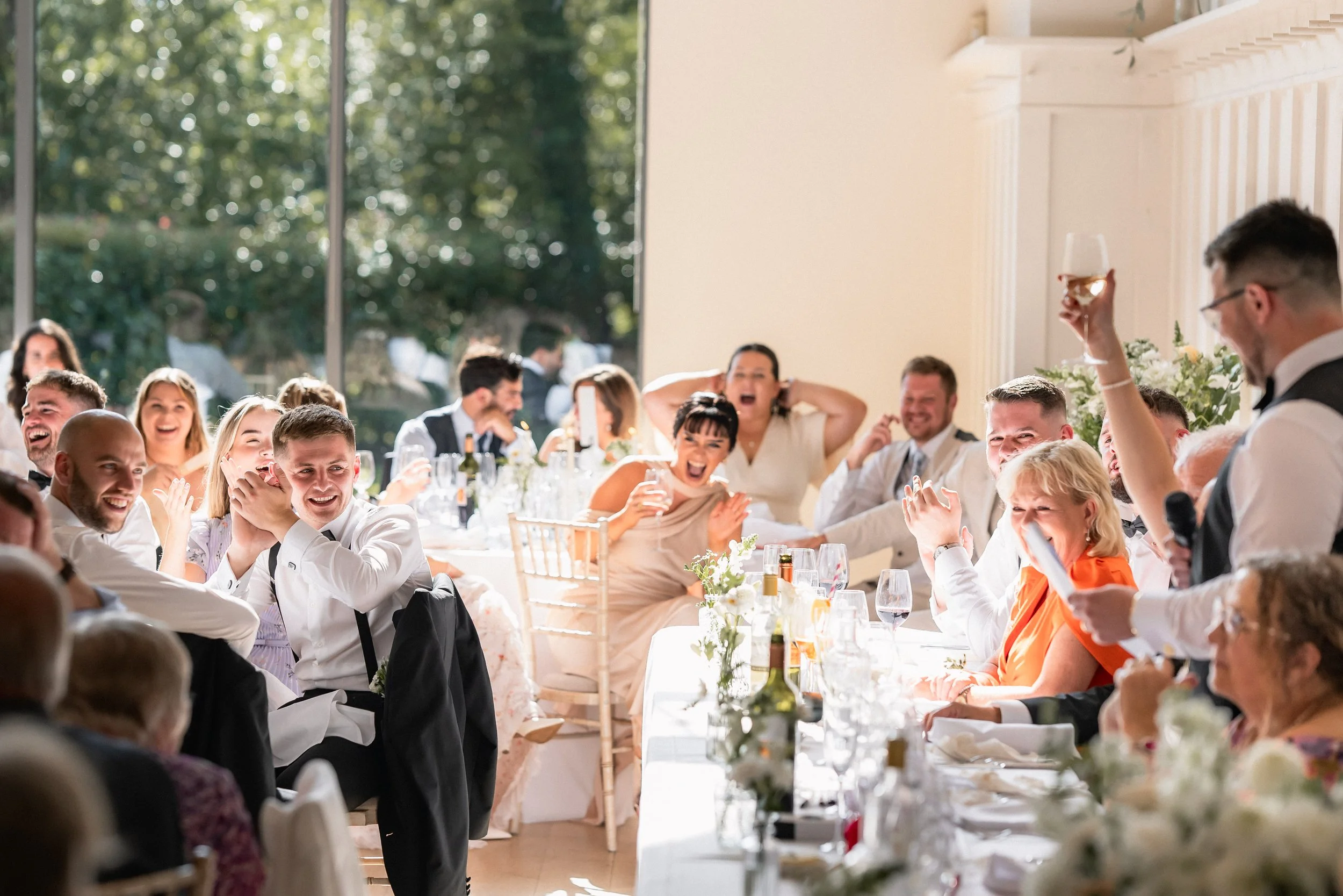 People at a wedding reception laughing and enjoying themselves, with a man giving a toast and a woman cheerfully reacting, inside a bright, decorated hall.