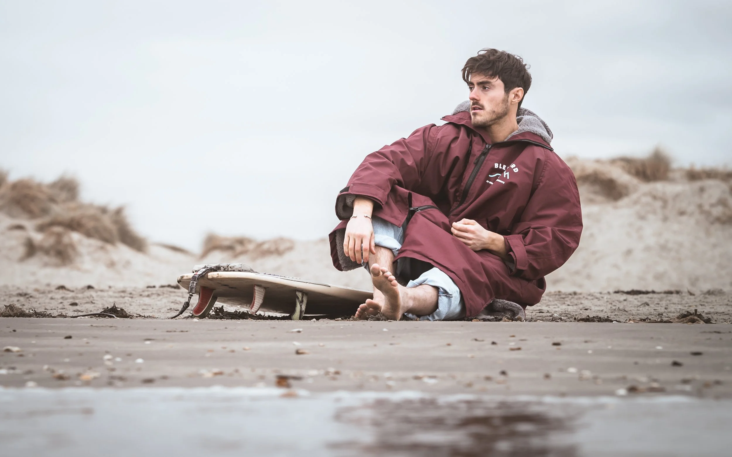 A young man sitting on a sandy beach after falling off his surfboard, with the surfboard lying on the sand nearby.
