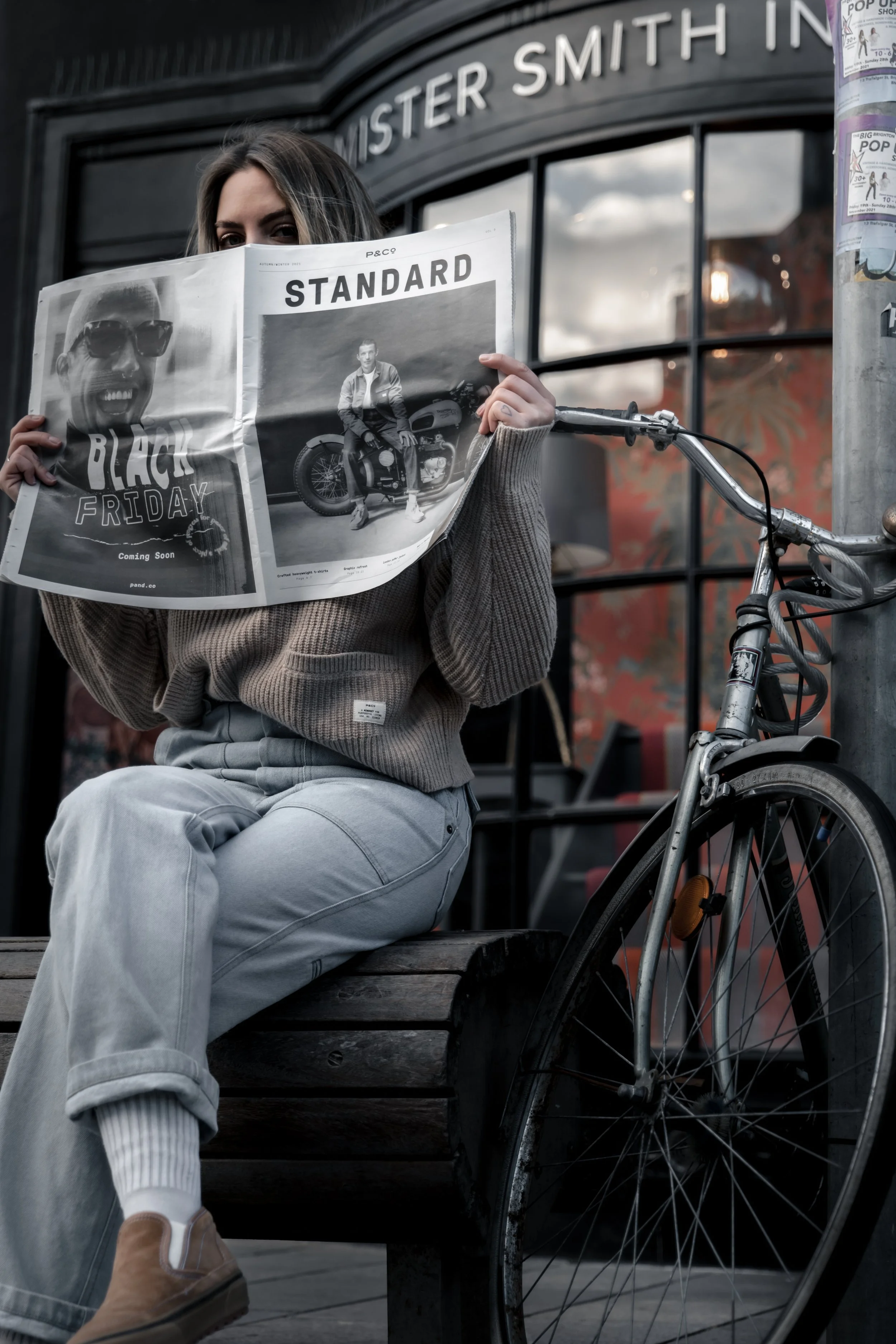 A woman sitting on a wooden bench, holding a newspaper in front of her face. The newspaper features a black-and-white photo of a man with sunglasses and text about Black Friday. There is a vintage bicycle parked beside her, and she is outside a shop 