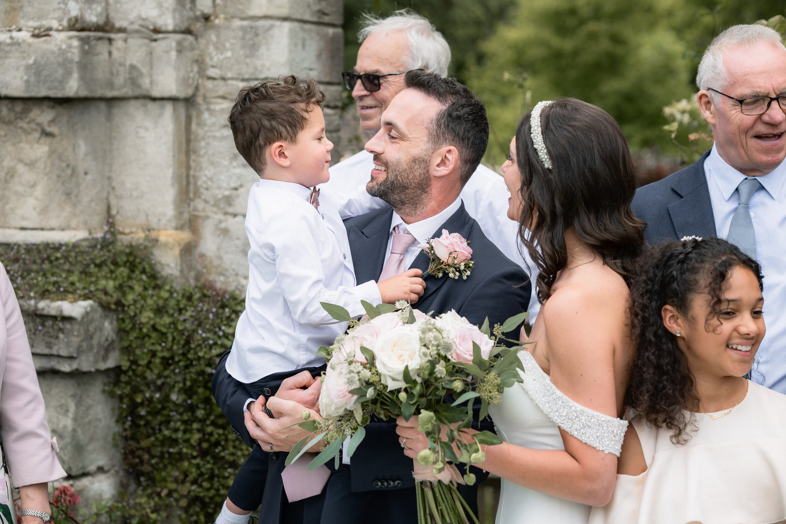 A wedding celebration outdoors with people smiling and interacting, including a man holding a young boy with a pink boutonniere, a woman in a white dress holding a bouquet, and guests in the background.