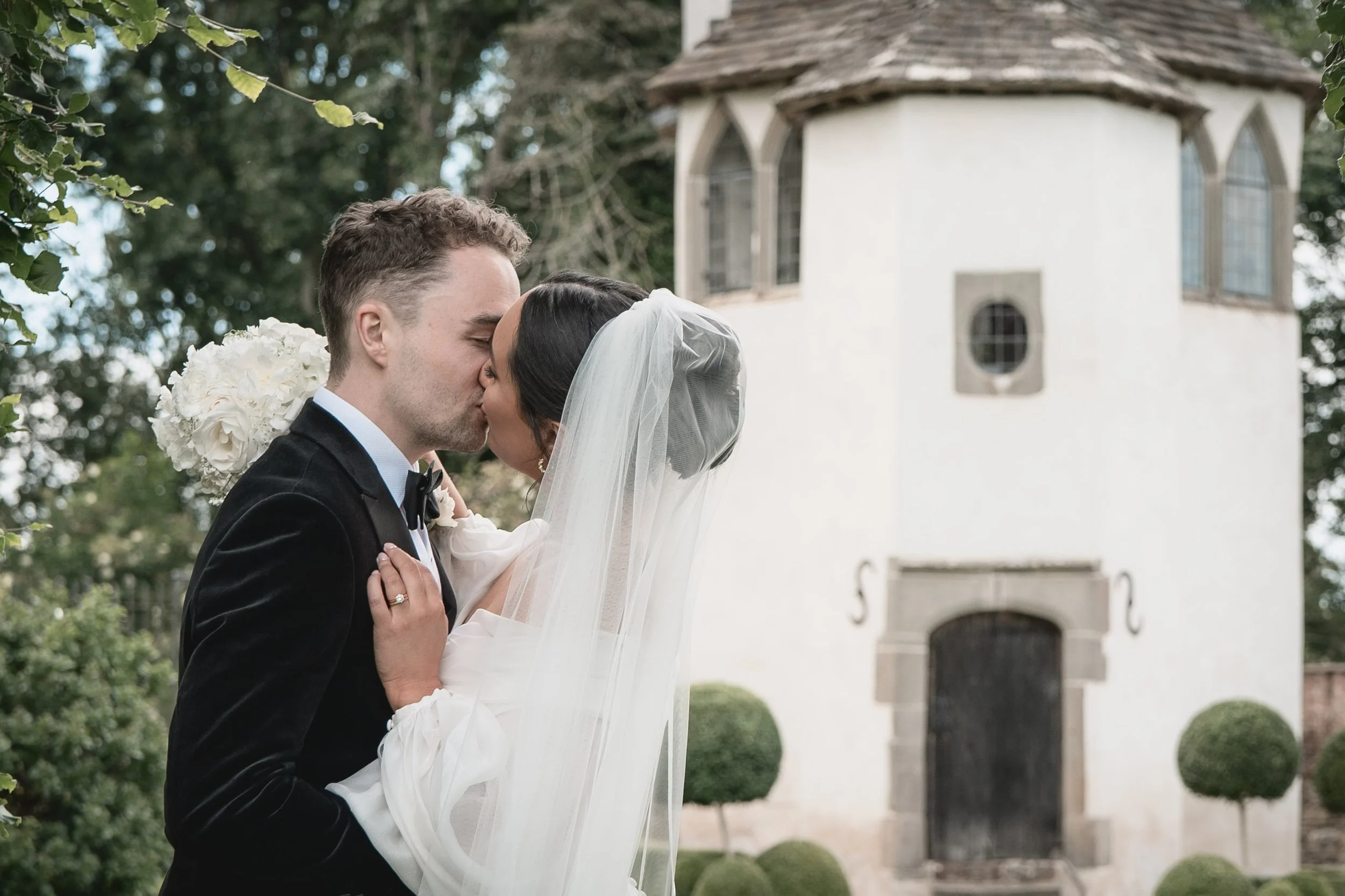 A newlywed couple sharing a kiss outdoors in front of a small white castle-like building, with surrounded trees and manicured bushes.