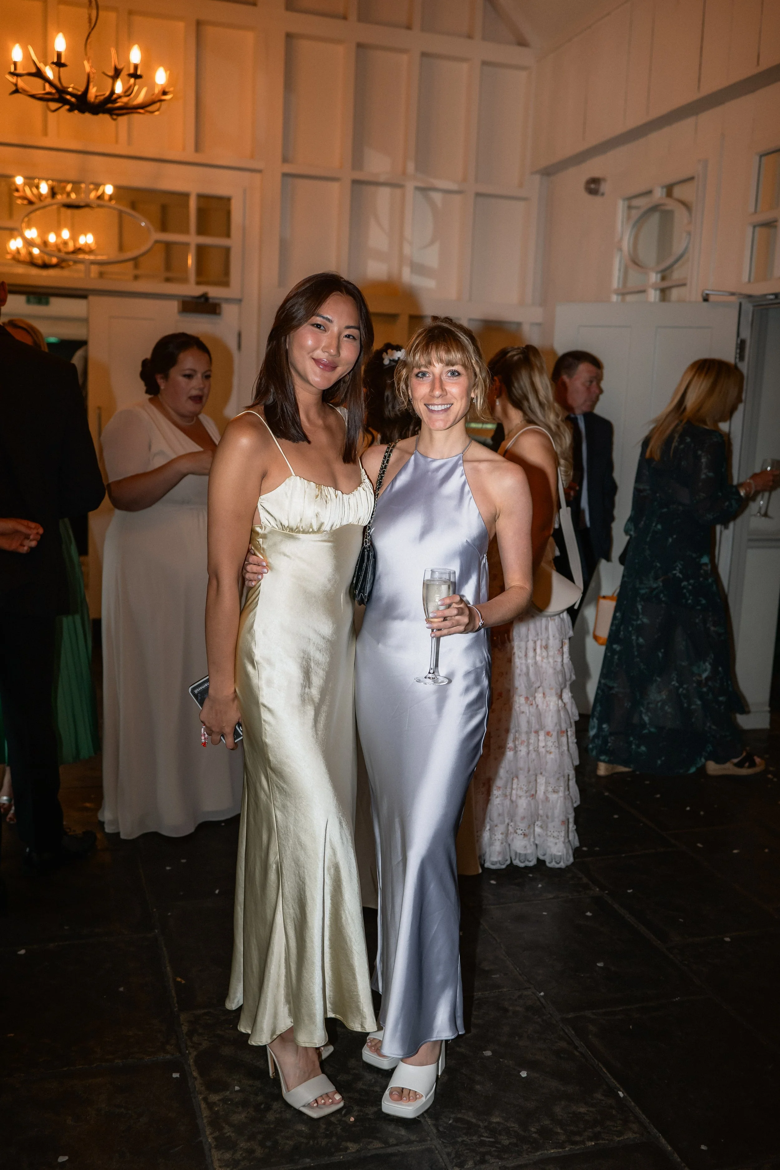 Two women in satin dresses posing at a formal event, one holding a glass of champagne, in a decorated indoor venue with chandeliers and other guests in the background.