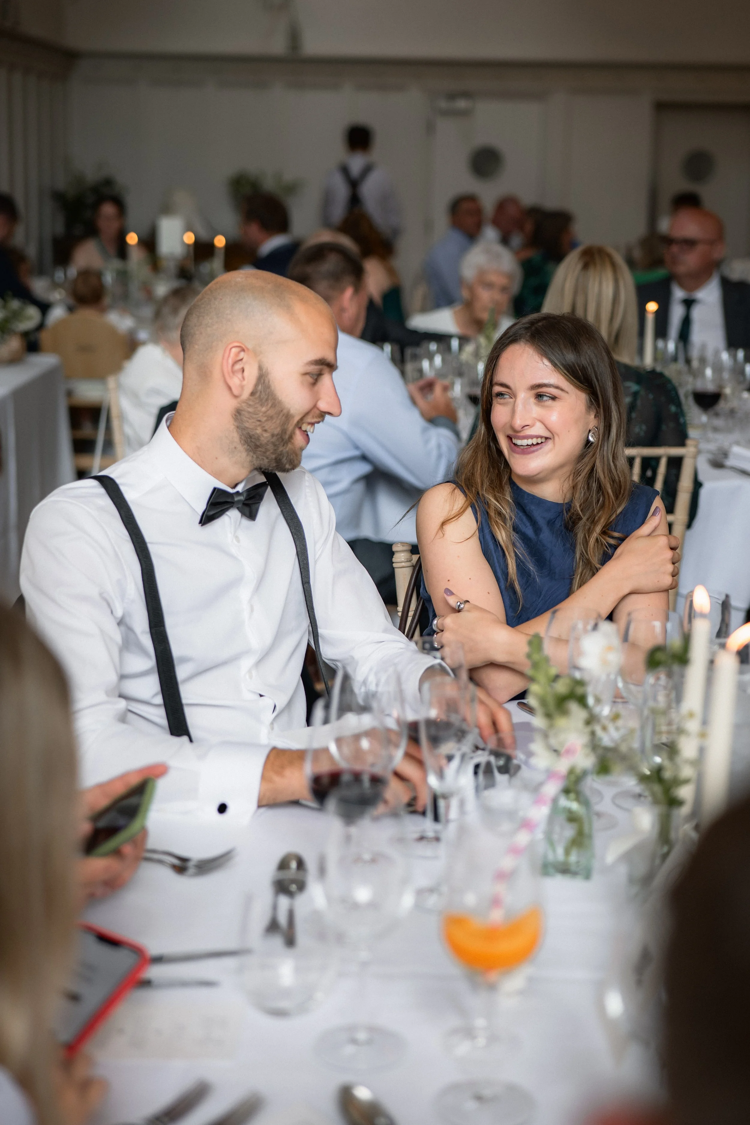 A man and a woman sitting at a formal dinner table, smiling and engaging in conversation at a wedding reception or similar event, with many other guests in the background.
