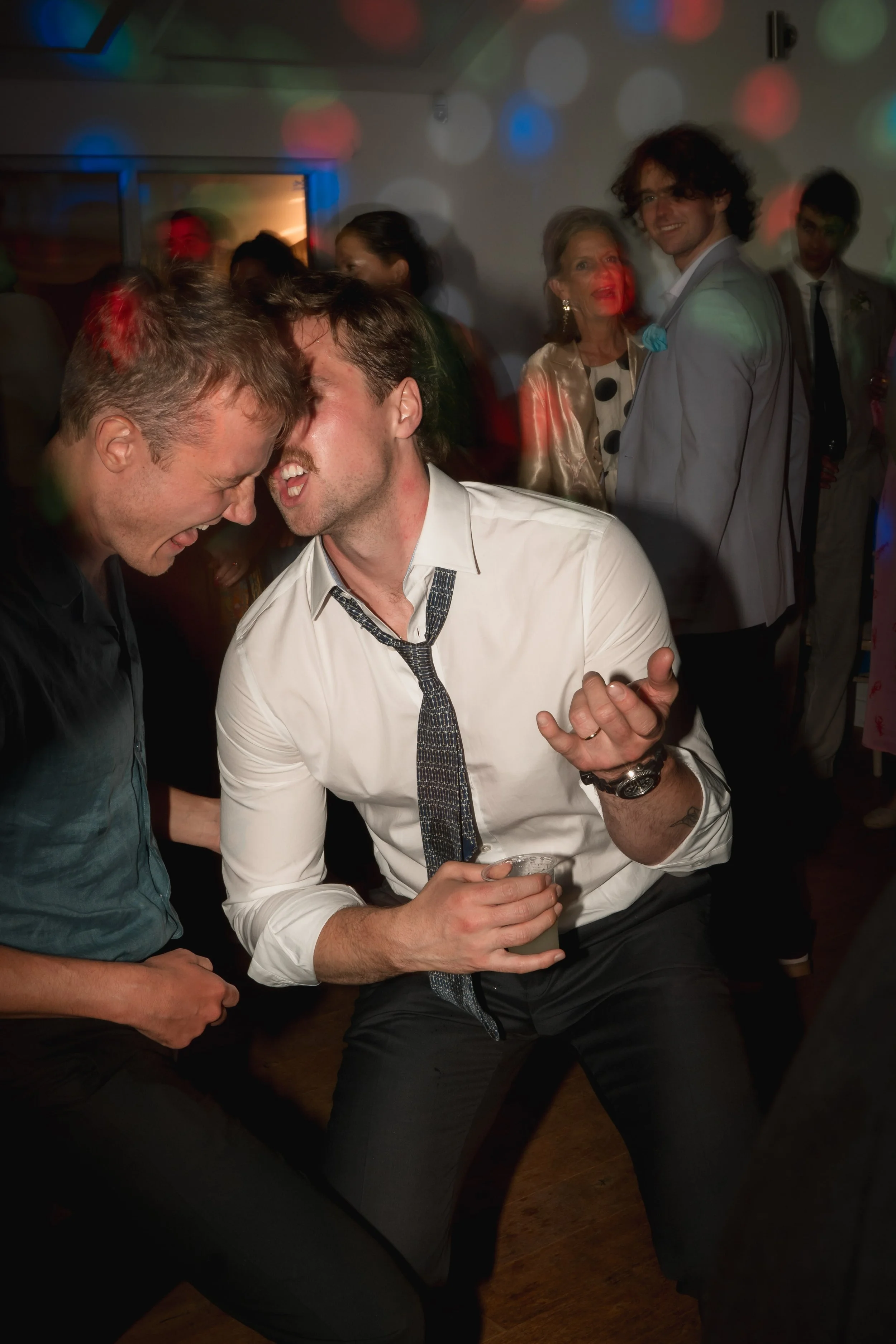 Two men dancing closely and smiling at each other at a lively party with colorful lights; other guests are visible in the background.