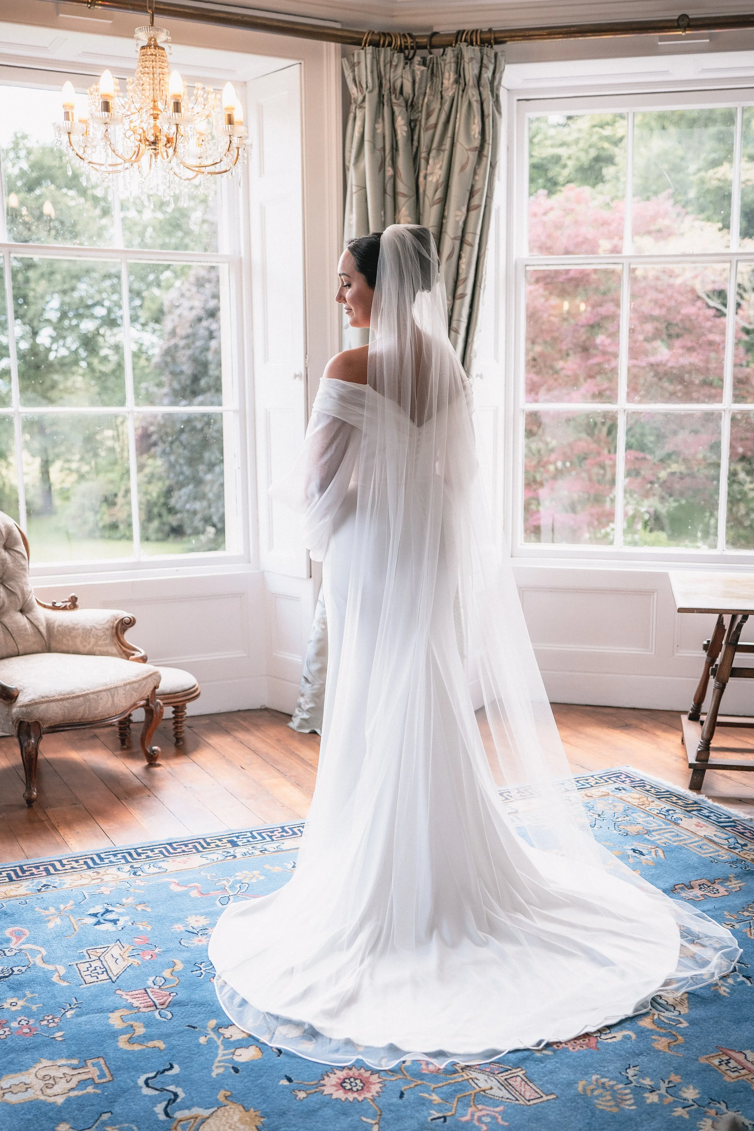 A bride in a white wedding gown and veil standing in front of large windows with a view of green trees and pink foliage.