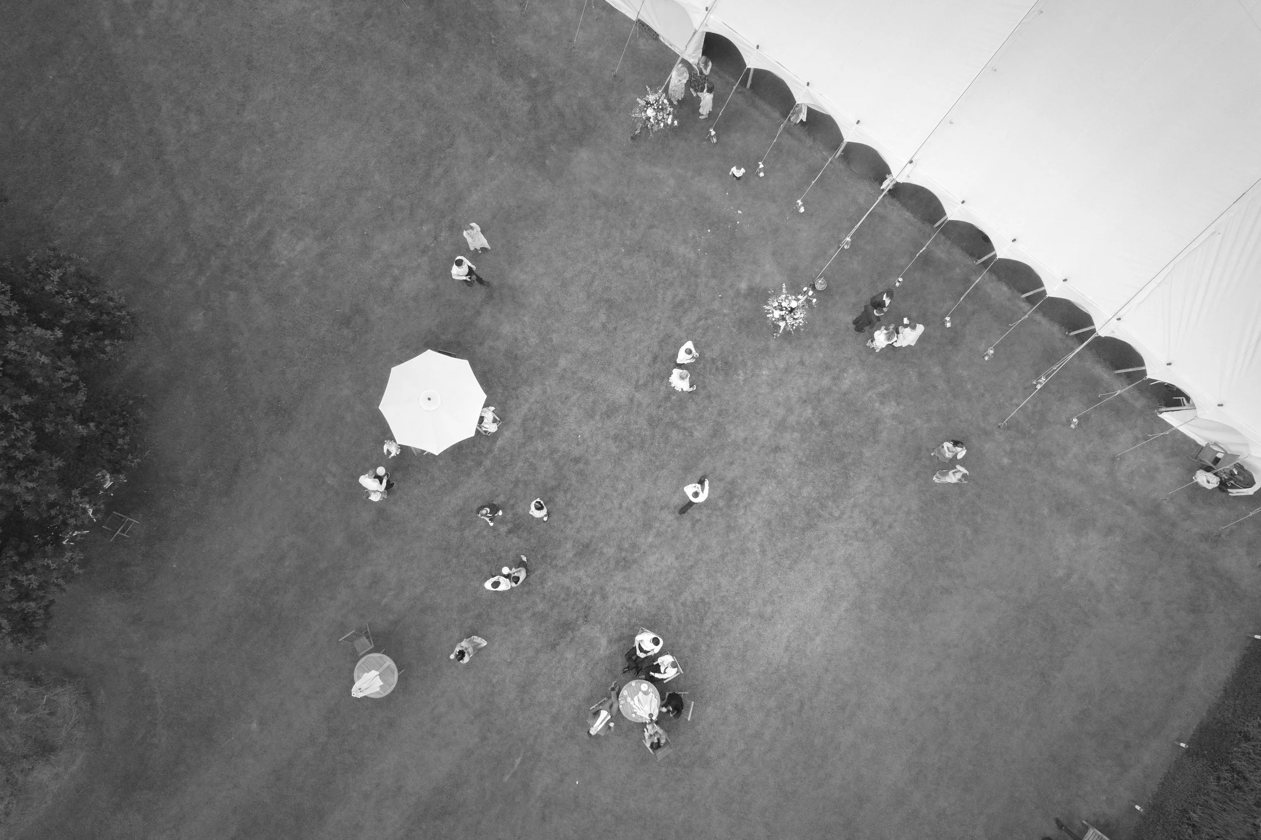 An aerial view of a wedding reception with guests gathered near a large white tent, some seated under an umbrella, and a few standing and chatting on a grassy area.