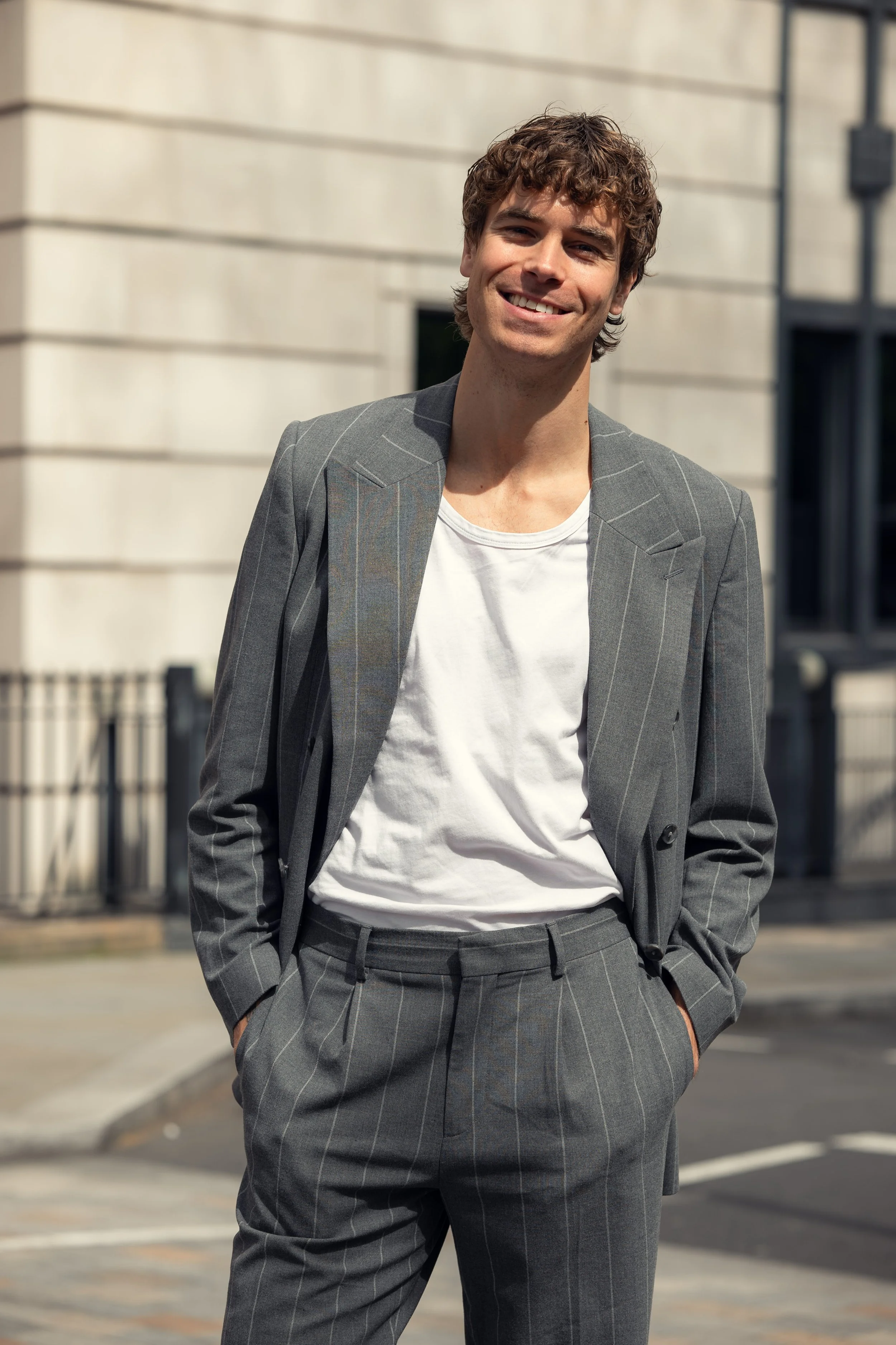 A young man in a gray pinstripe suit, white t-shirt, standing outdoors on a city street, smiling with hands in pockets.