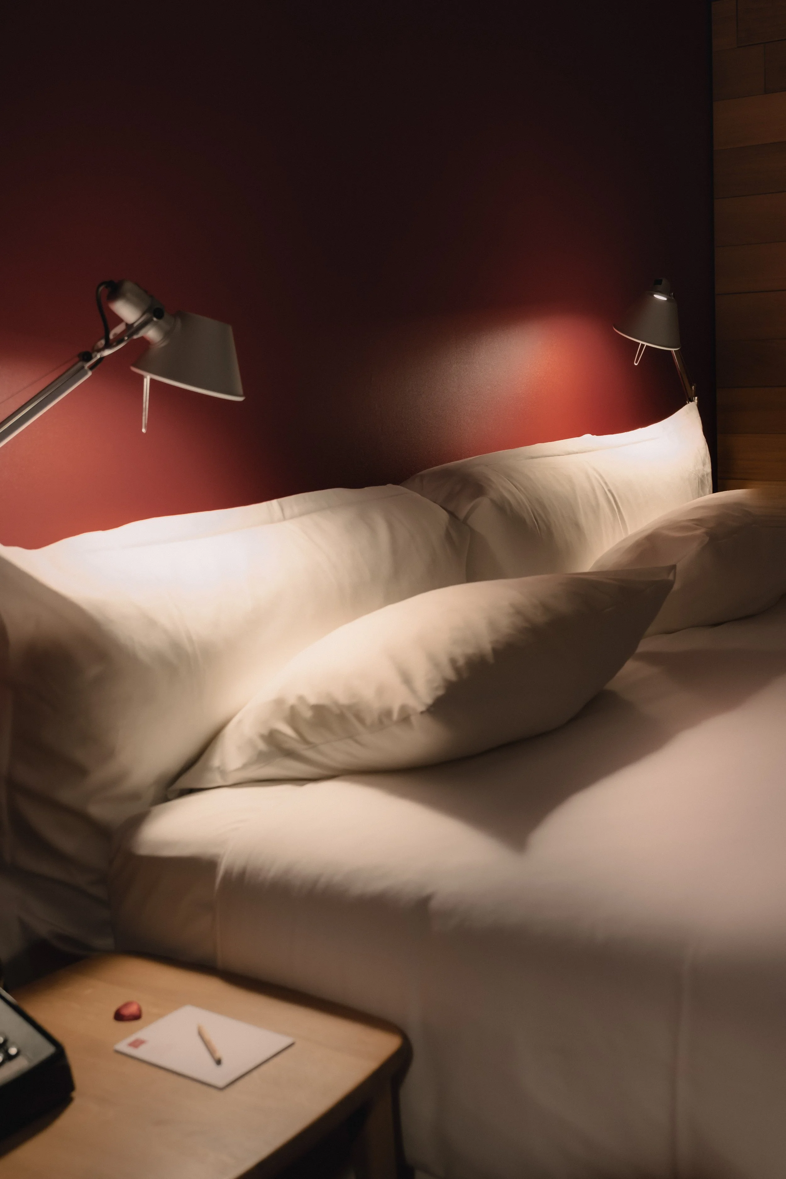 a bed with multiple pillows, bedside table with a notepad and pencil, wall-mounted reading lamps, and a dark red wall with wood paneling on the side