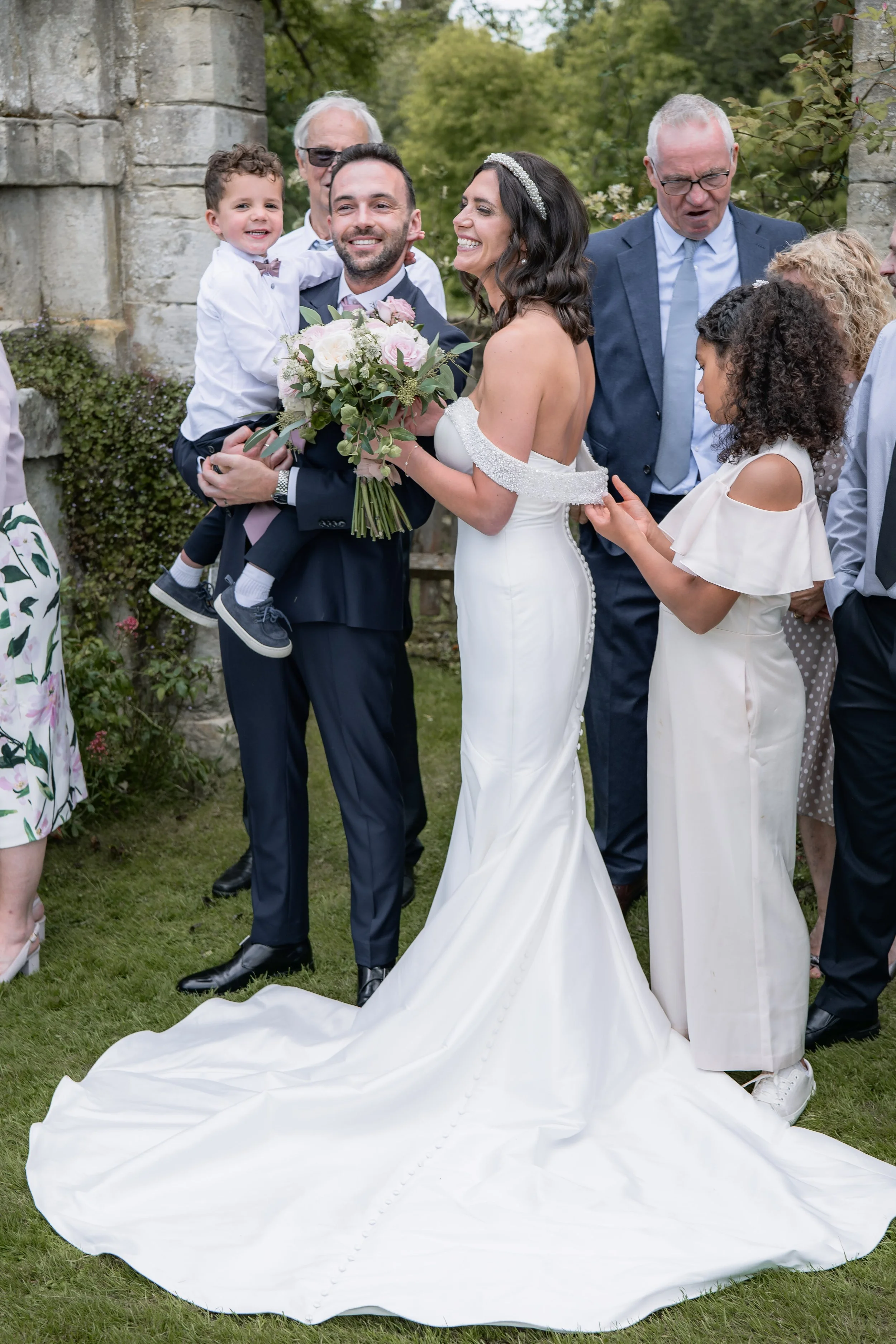 A bride in a white wedding gown and a groom in a tuxedo holding a young boy at a wedding ceremony outdoors, surrounded by family members.