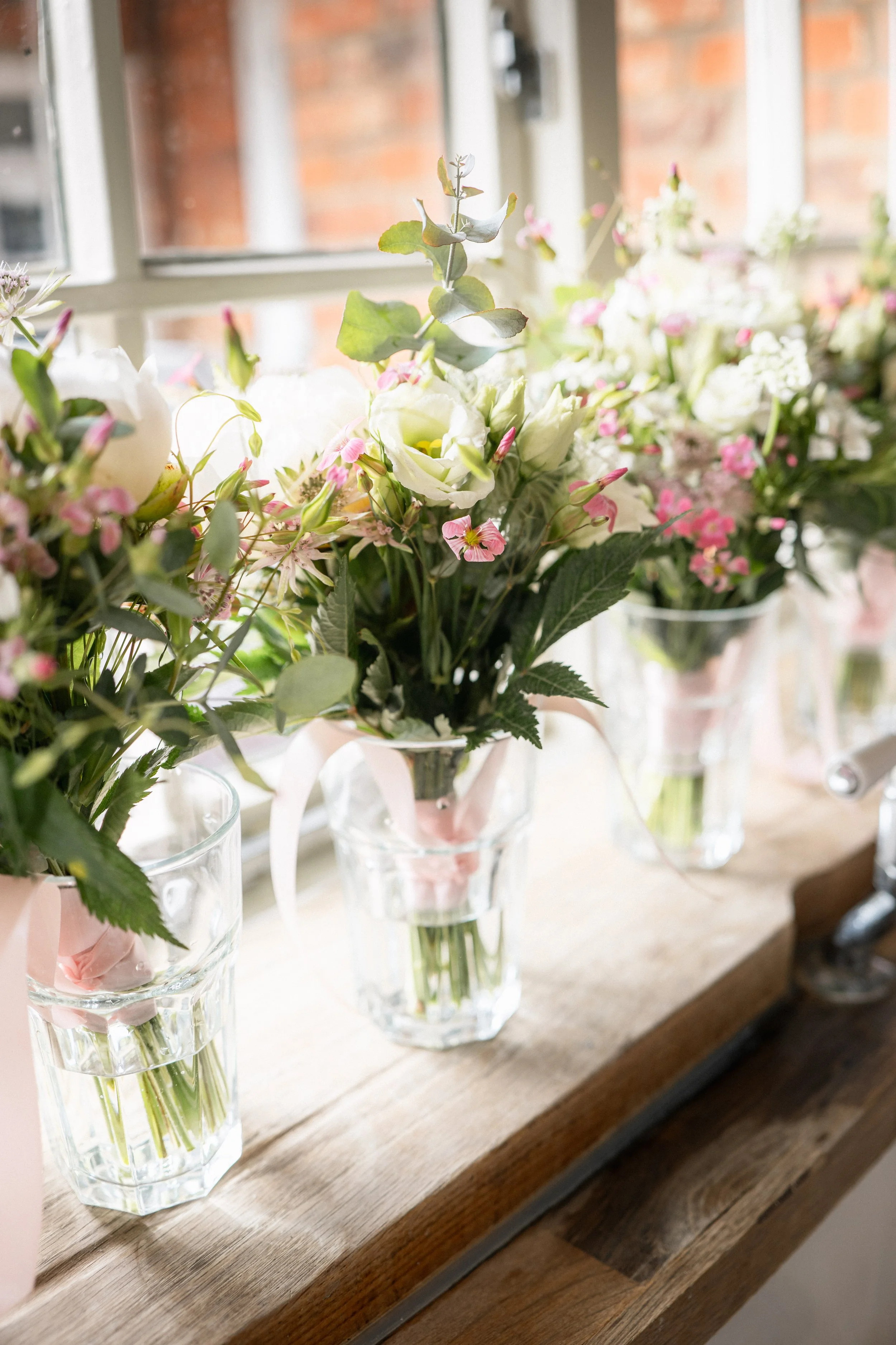 Vases with fresh pink and white flowers on a wooden shelf near a window.