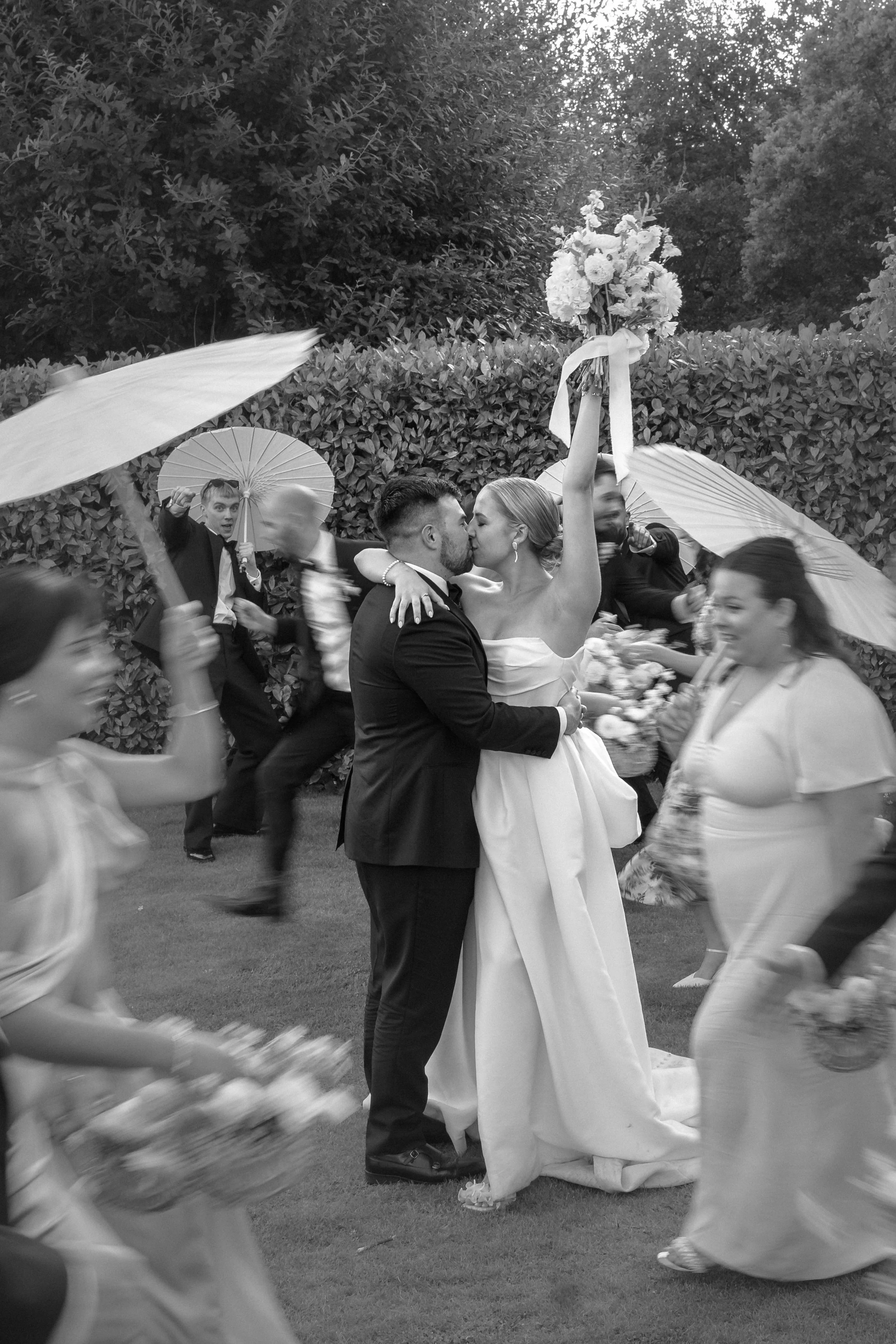 Black and white photo of a wedding celebration with a couple kissing, the bride holding a bouquet with ribbons raised, guests with umbrellas and flowers surrounding them.