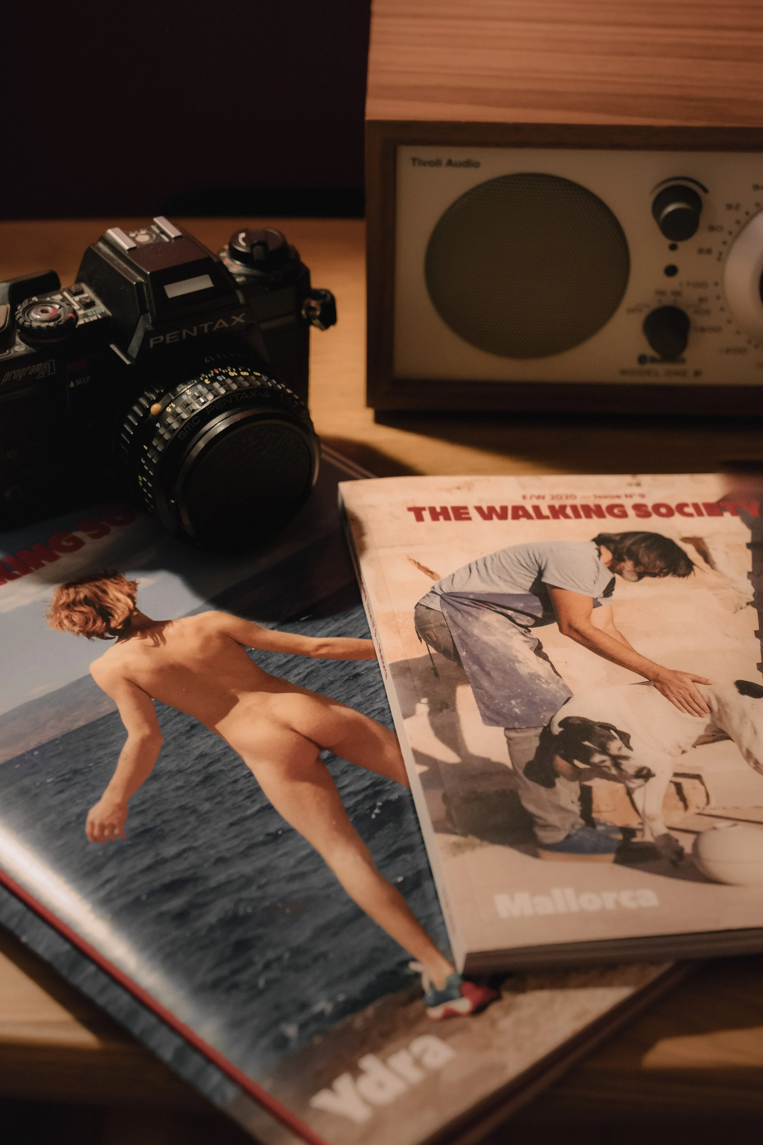A vintage camera, a small wooden radio, and a magazine titled 'The Walking Society Mallorca' are placed on a wooden surface. The magazine features an image of a man and a dog, and a photograph of a woman standing on a beach.