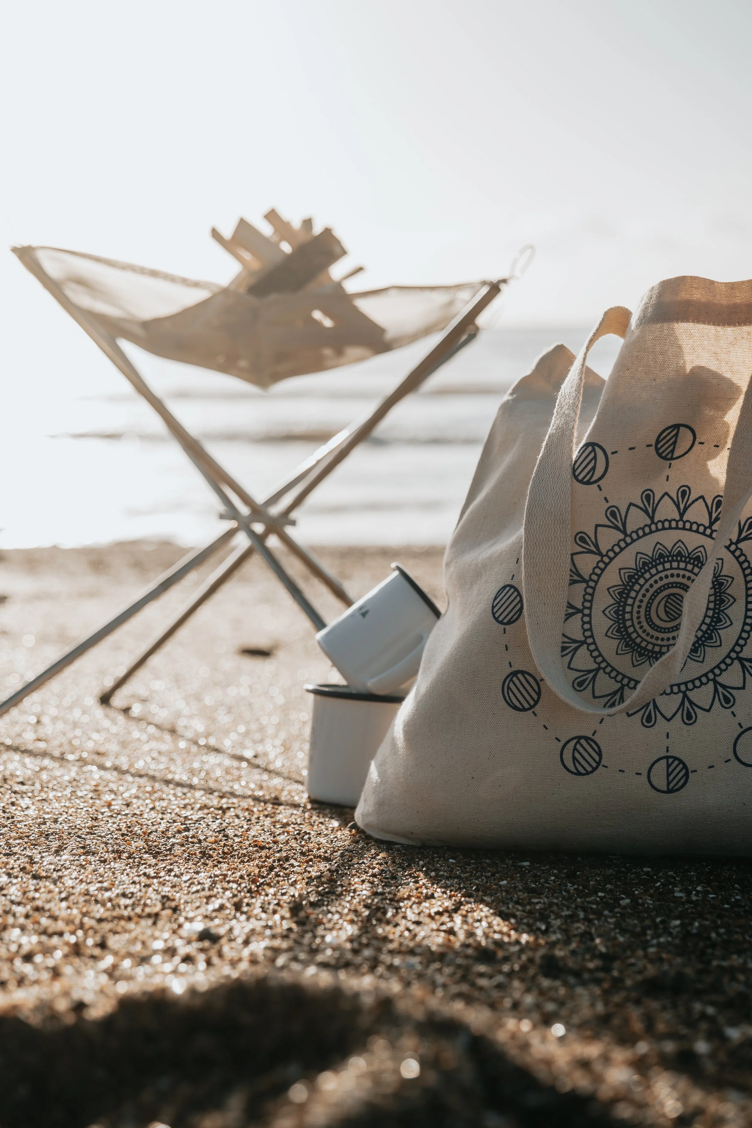 Beach scene with a tote bag, a mug, and a foldable beach chair near the shoreline.