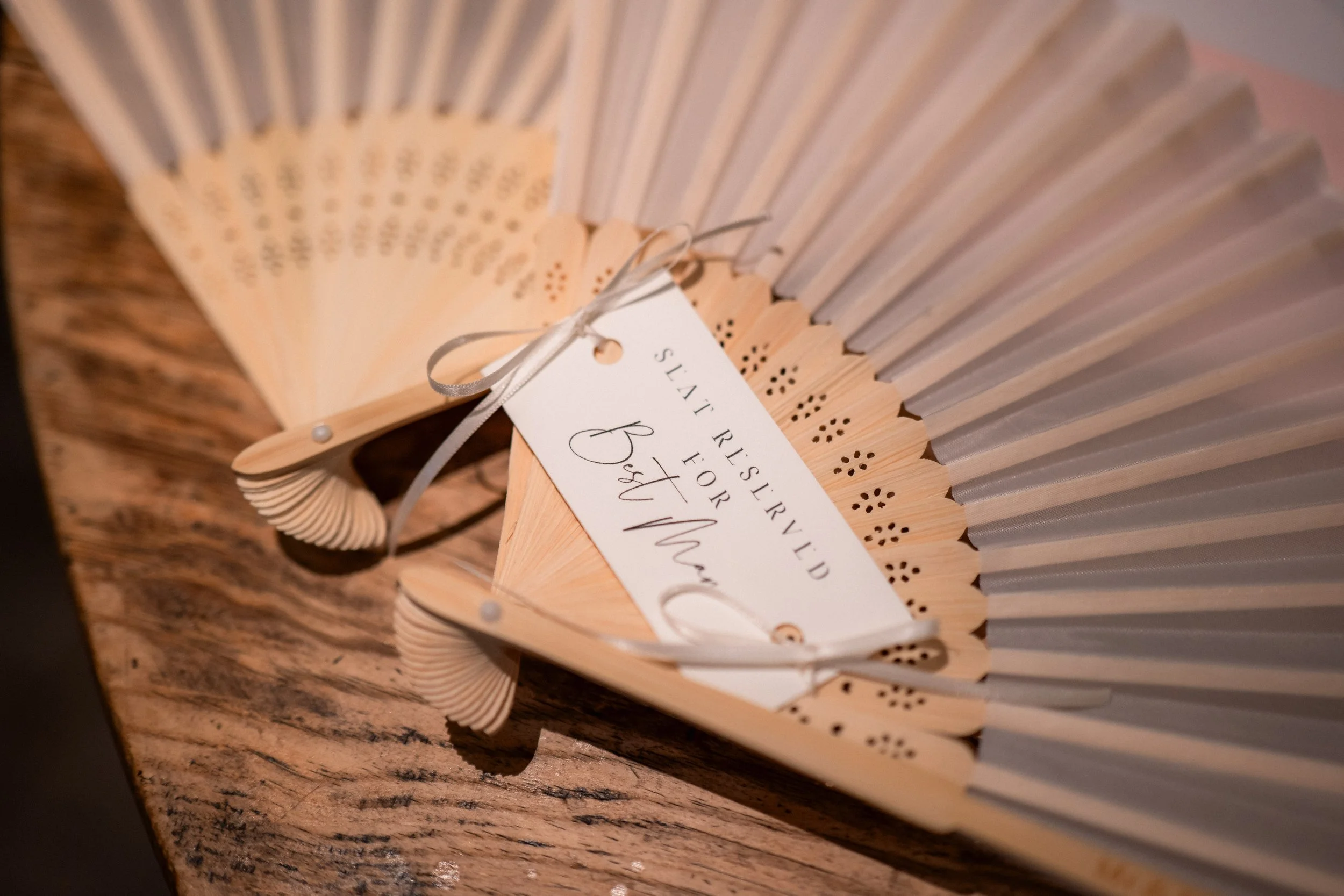 A decorative wooden hand fan with a tag reading "seaf preserve for best mom" is placed on a wooden surface.