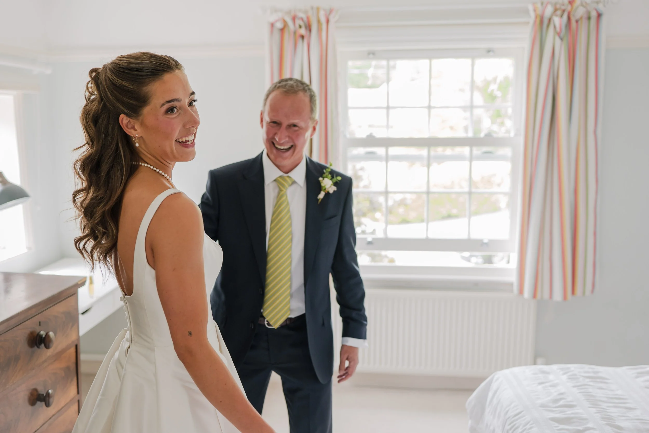 A bride and an older man, possibly her father, sharing a joyful moment in a bedroom with a window and curtains in the background.