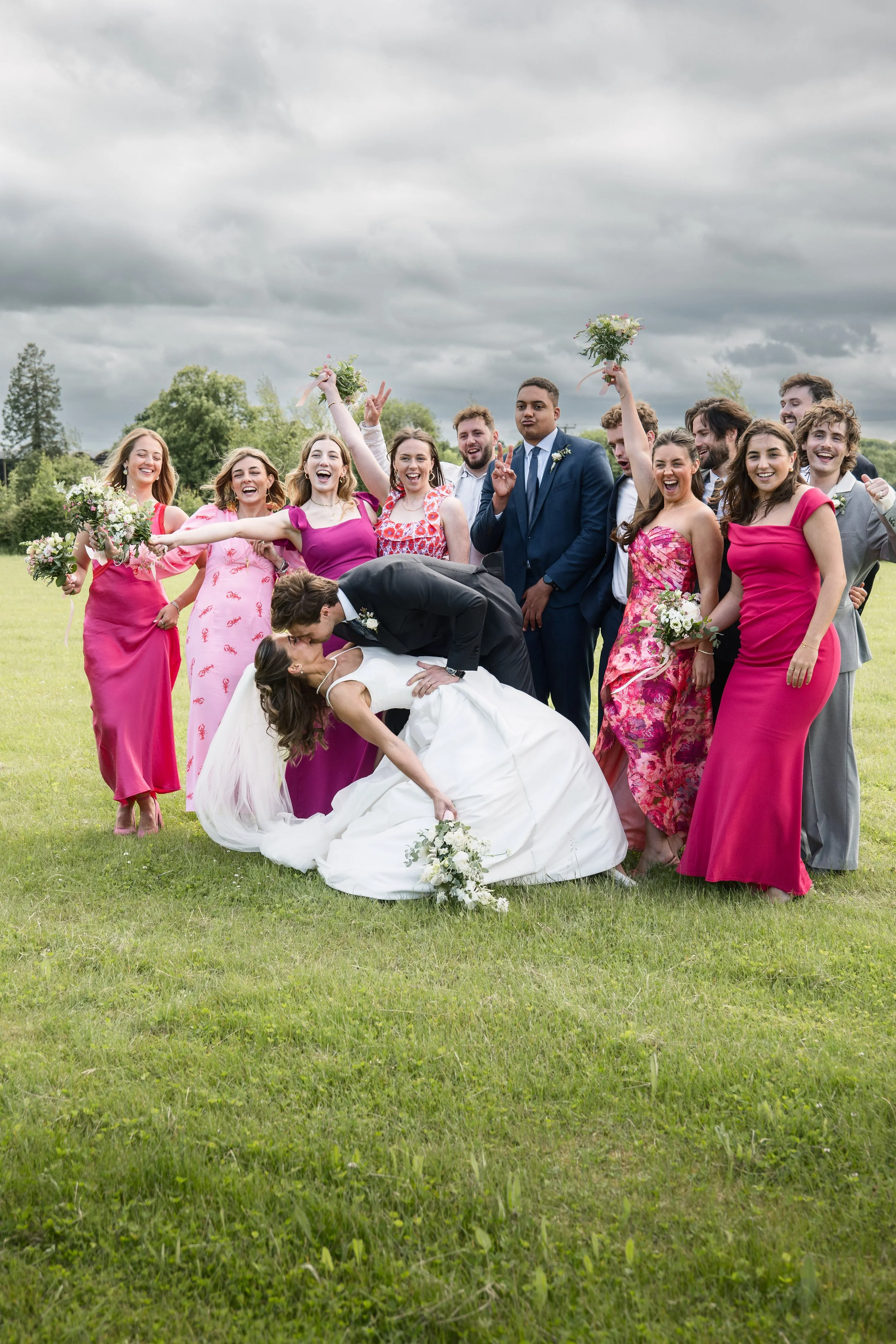 A group of wedding guests celebrating outdoors on a grassy field, with a bride and groom in the center kissing. The guests are wearing colorful dresses and suits, smiling, with some holding bouquets. The sky is overcast with dark clouds.