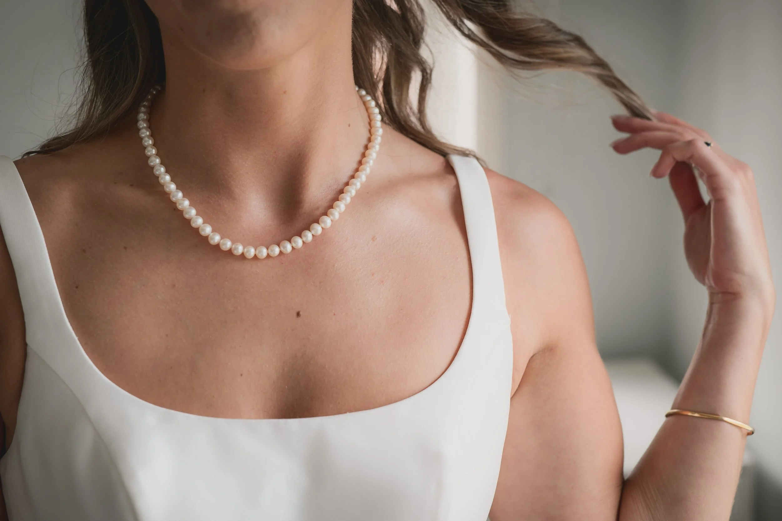 Close-up of a woman wearing a white sleeveless top, pearl necklace, gold bracelet, and a ring, with her hand near her hair.