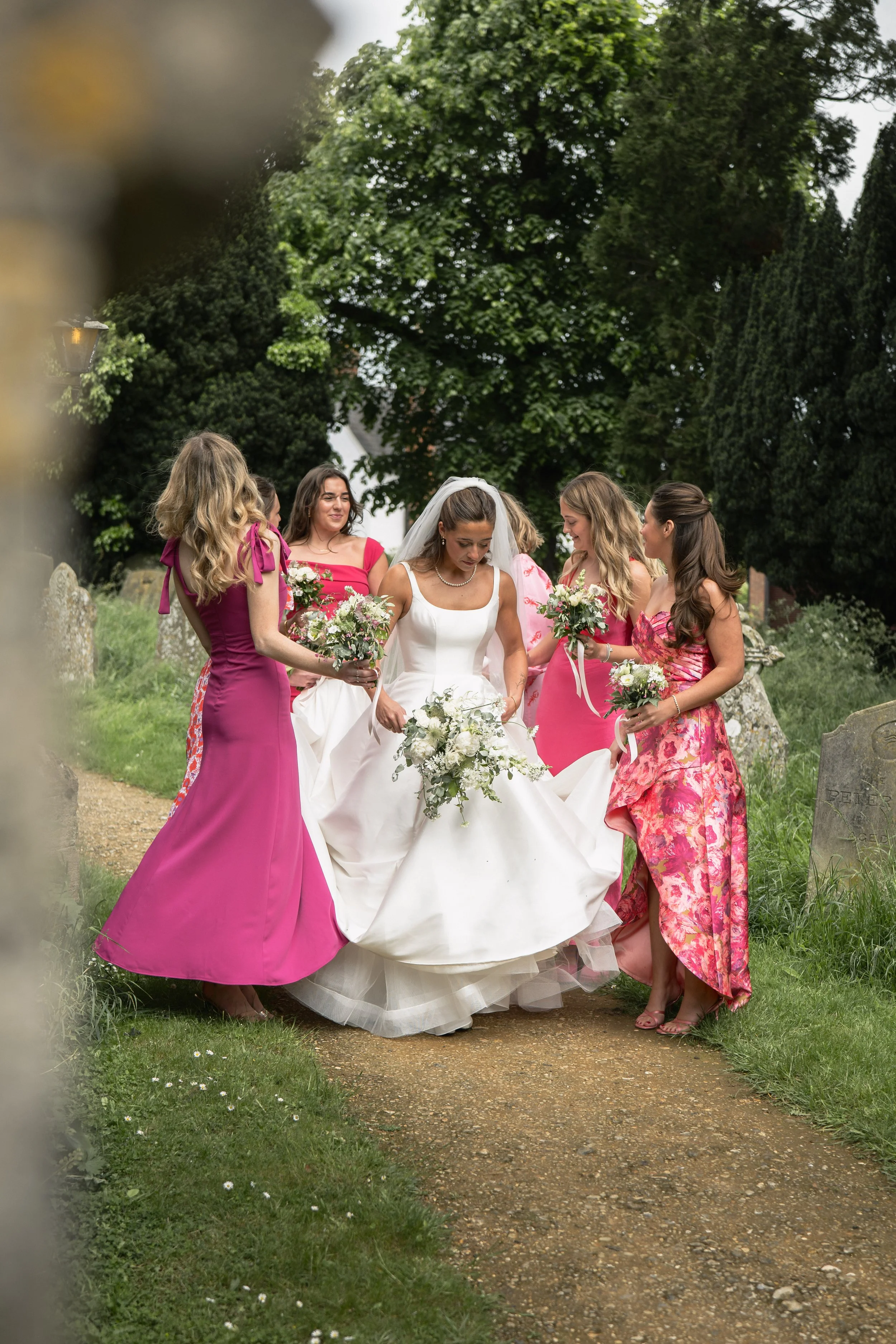 Bride in a white wedding dress with veil and bouquet walking on a garden path, surrounded by four women in colorful dresses holding bouquets, in a lush green outdoor setting.