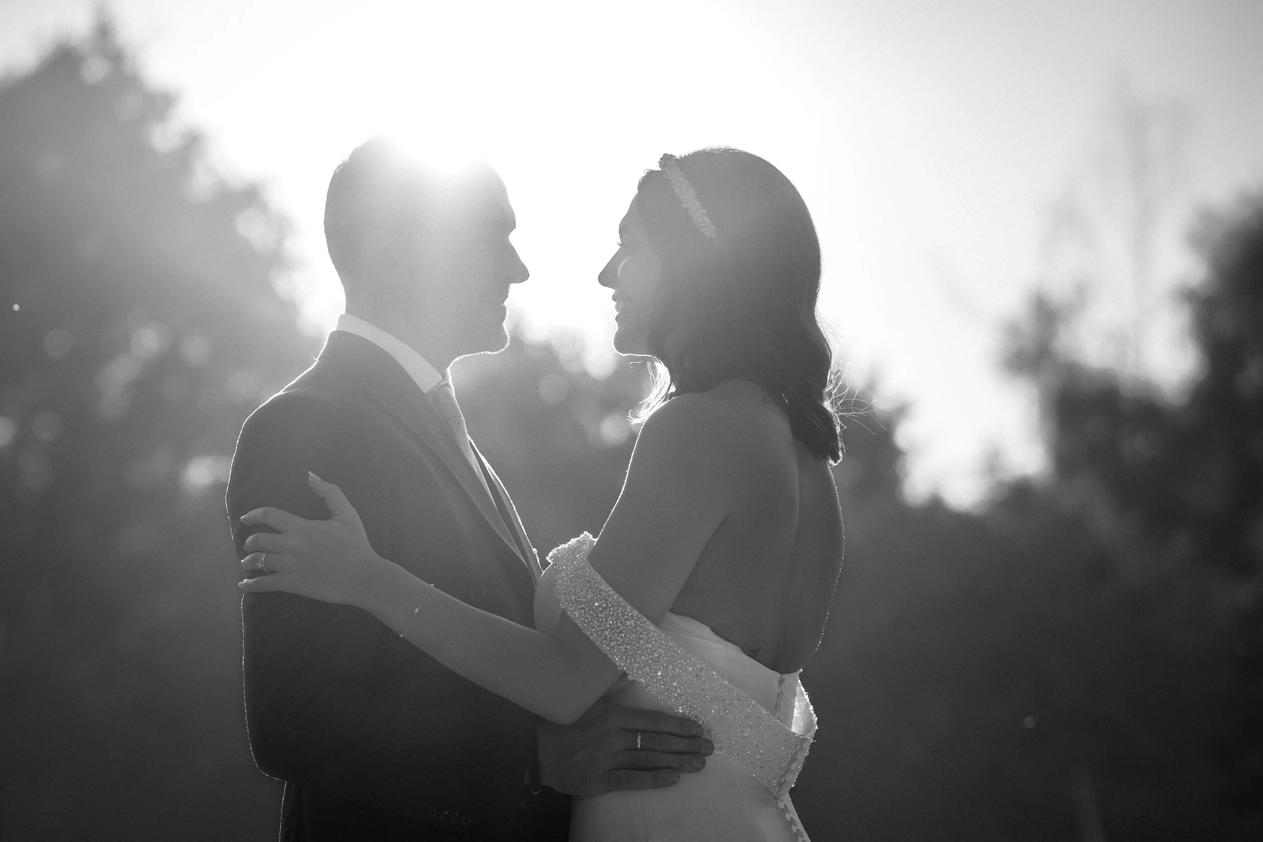 A couple in wedding attire stands close together outdoors, holding each other and looking into each other's eyes with the sun setting behind them.