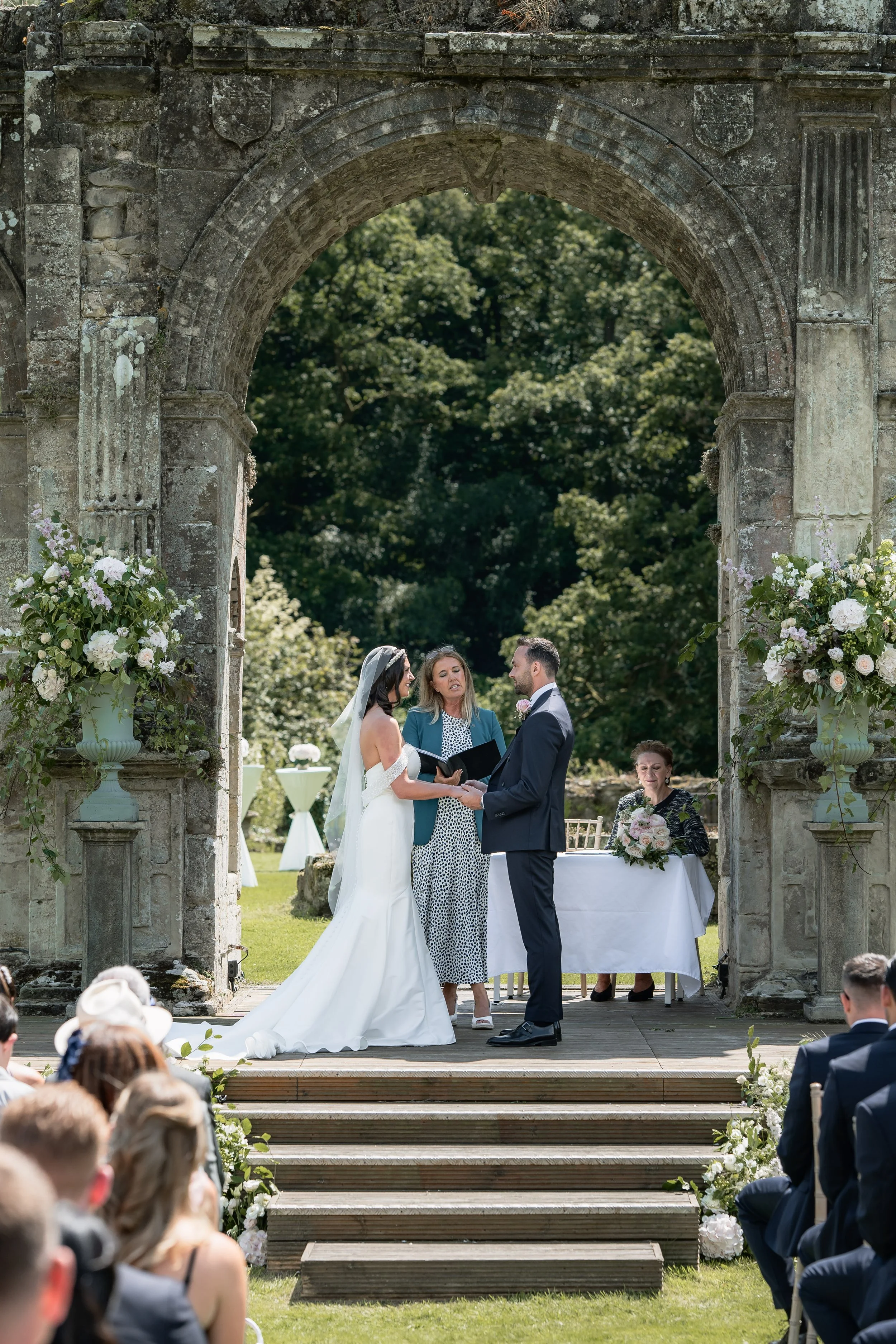A wedding ceremony taking place outdoors under an archway with stone pillars, with a bride and groom holding hands and exchanging vows. The bride wears a white gown, and the groom wears a dark suit. An officiant stands between them, and a woman holdi