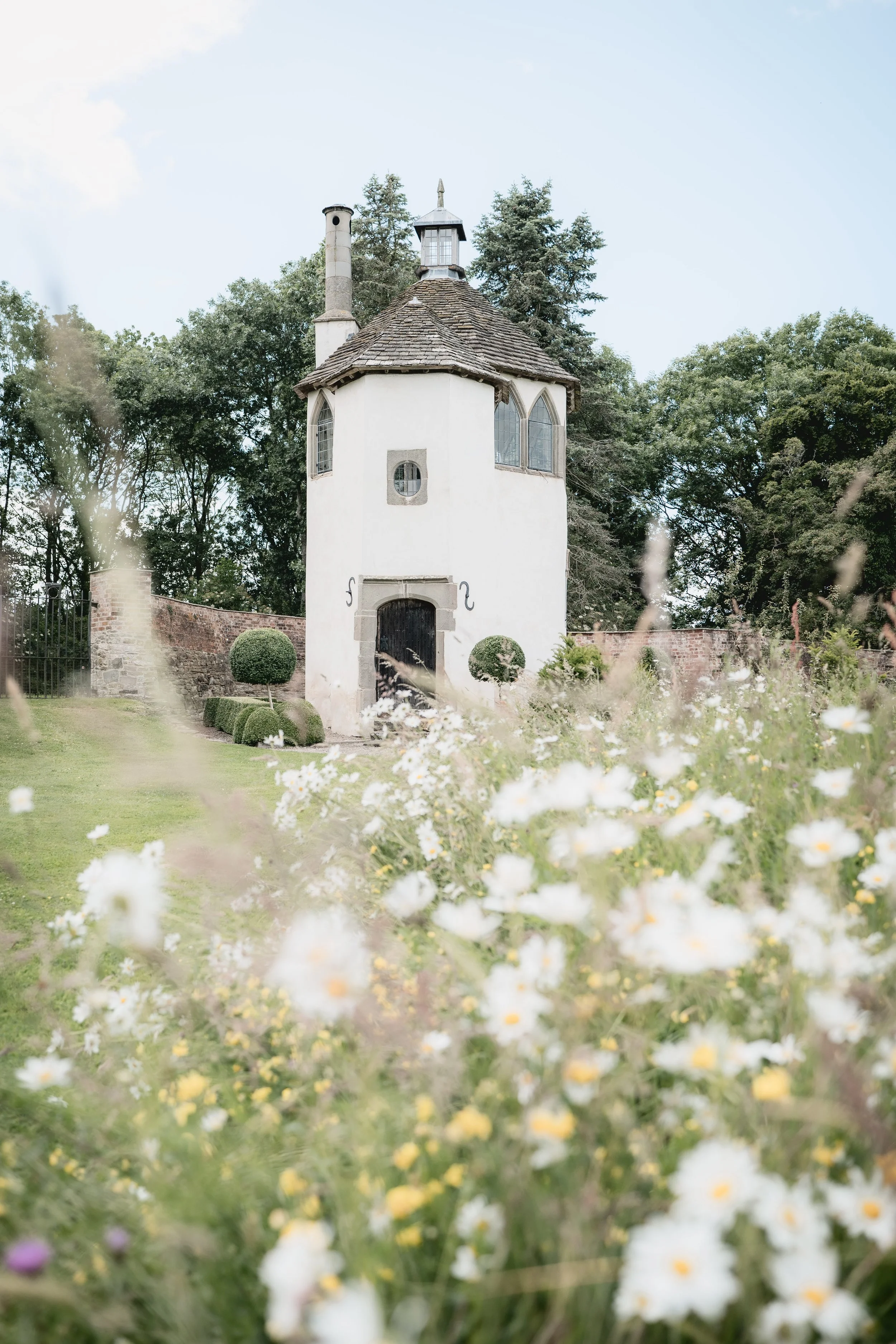 A small white tower with a thatched roof and a weather vane surrounded by a well-kept garden and trees.