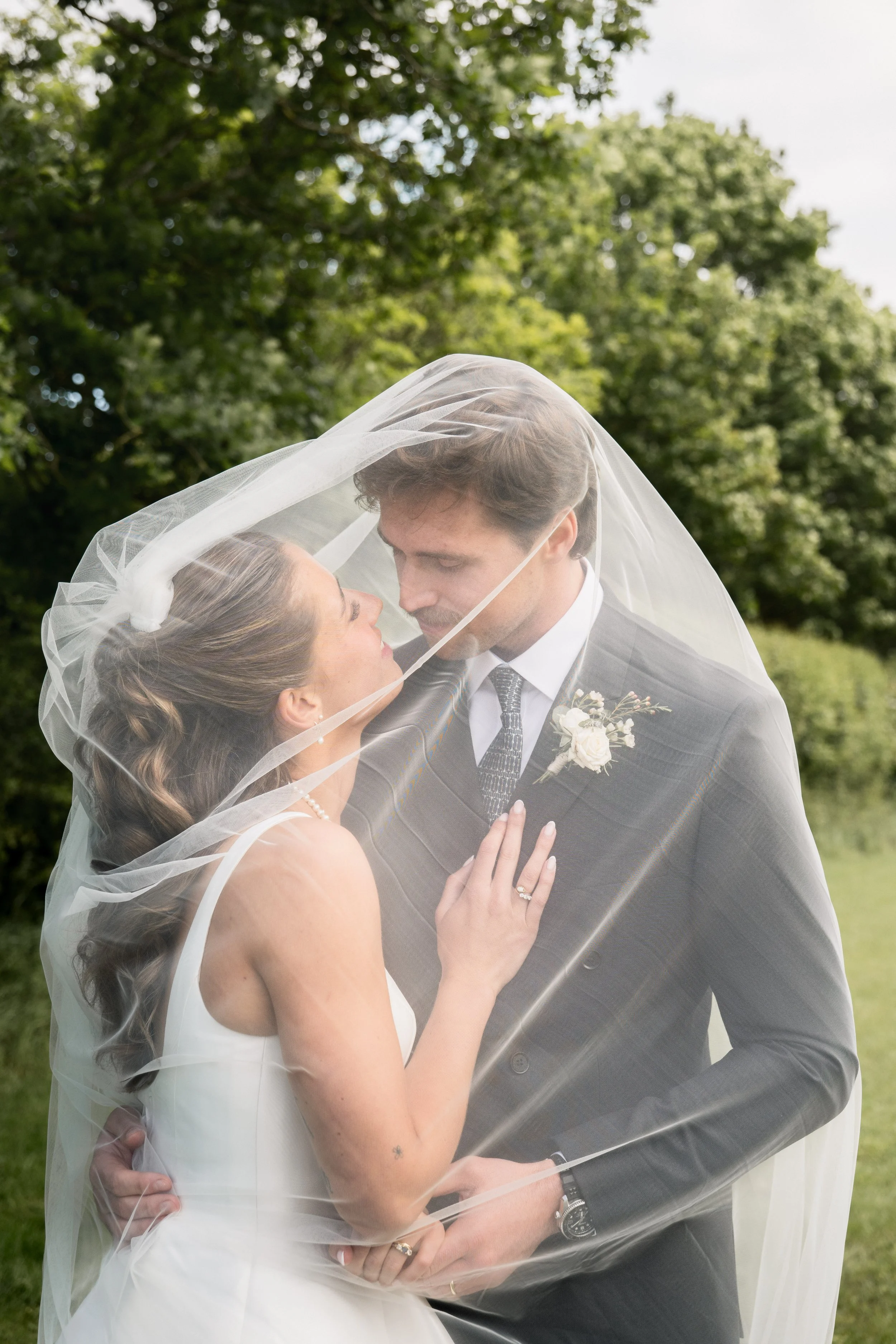 A bride and groom posing outdoors for a wedding photo, wrapped together in a sheer veil under a cloudy sky with green trees in the background.
