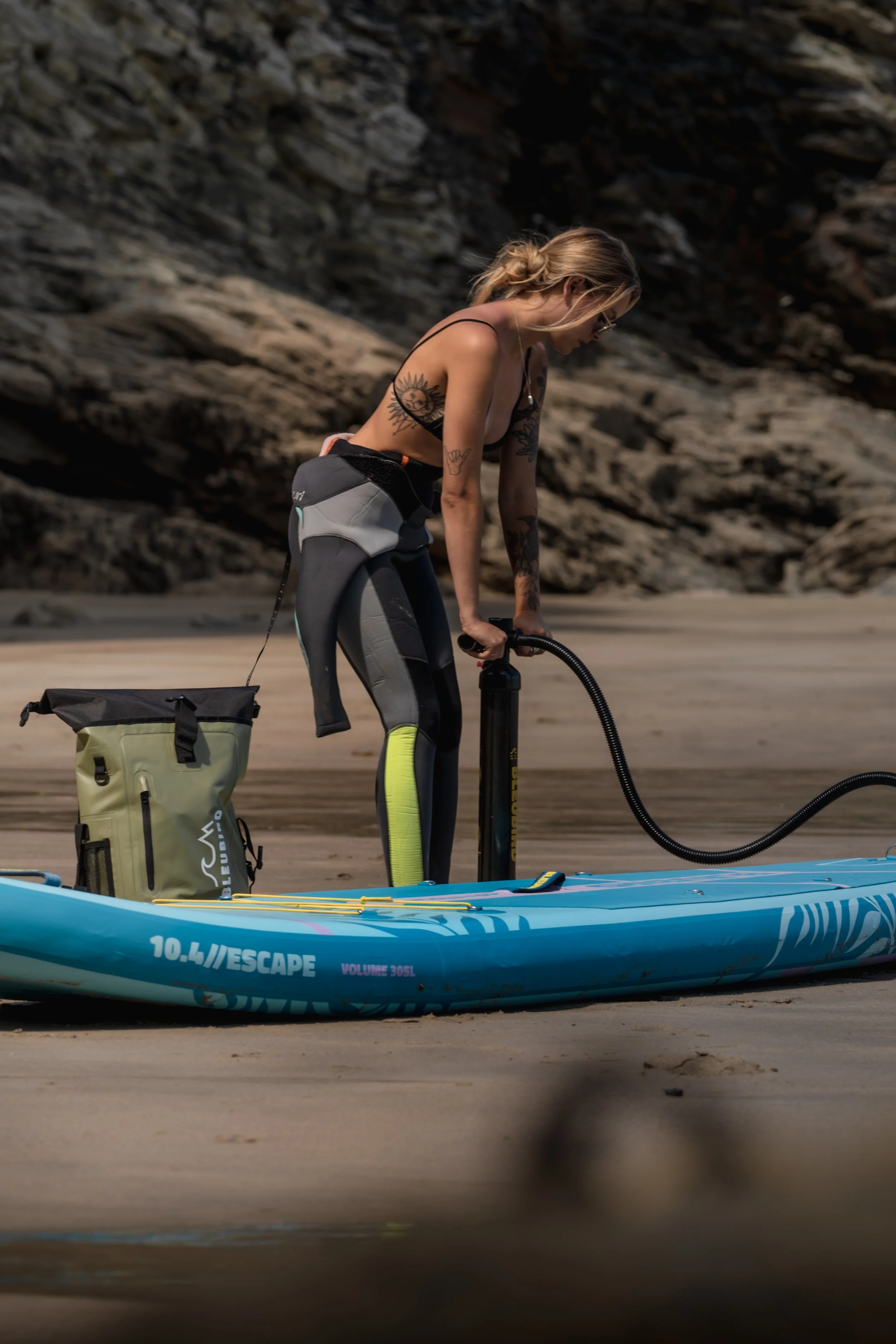 A woman in athletic gear blowing up an inflatable paddleboard on a sandy beach with rocky cliffs in the background.