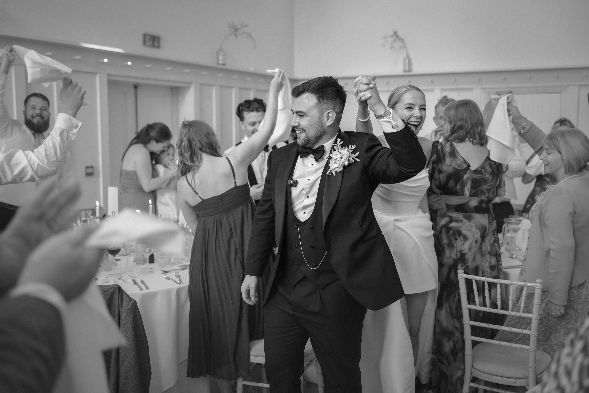 A wedding reception with the groom and bride dancing, surrounded by guests. The groom wears a tuxedo and the bride wears a white wedding dress. Guests are smiling, raising their hands, and enjoying the celebration.