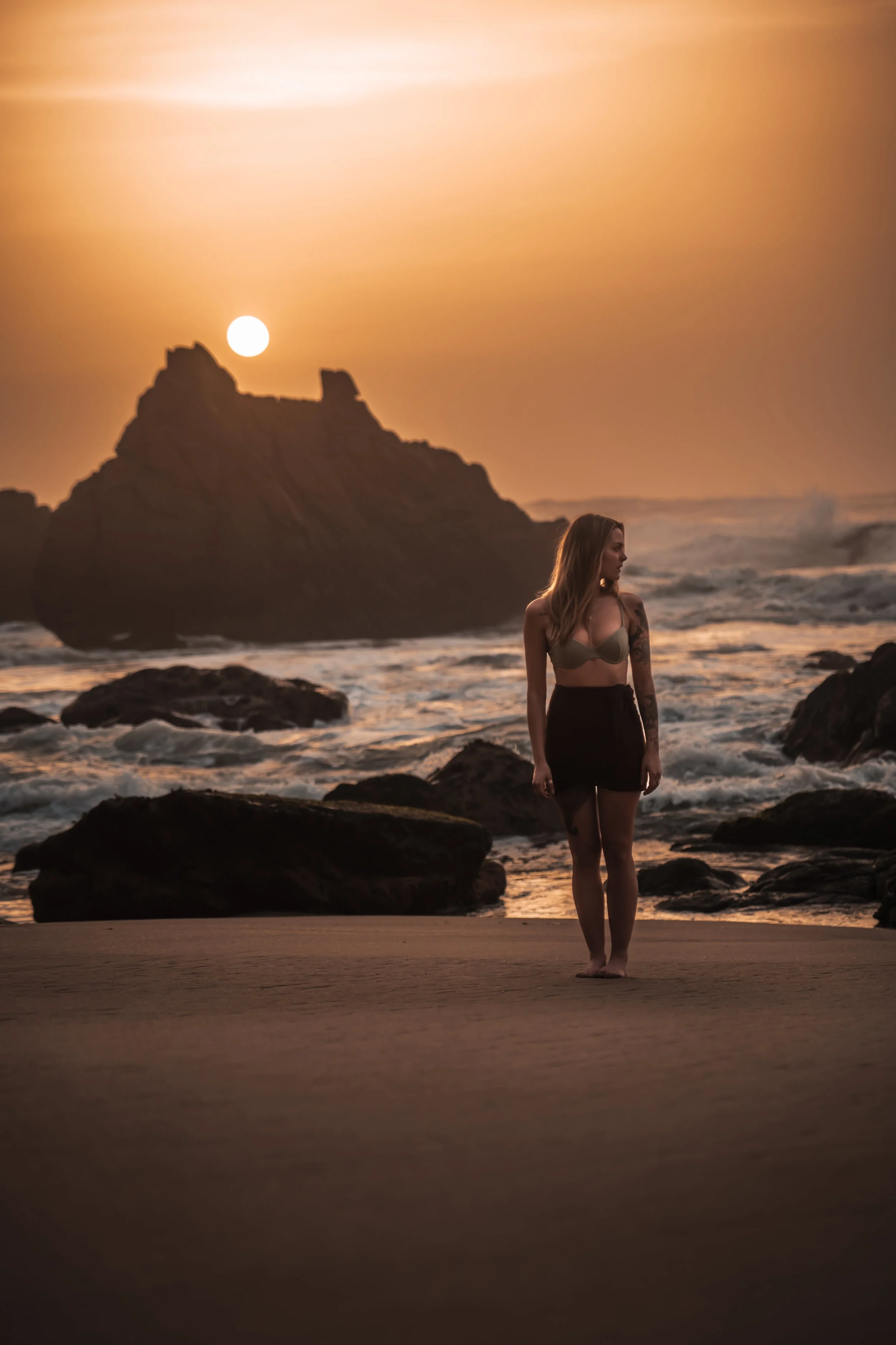 A woman with tattoos in a bikini top and skirt standing on the beach at sunset, with rocks and waves in the background.