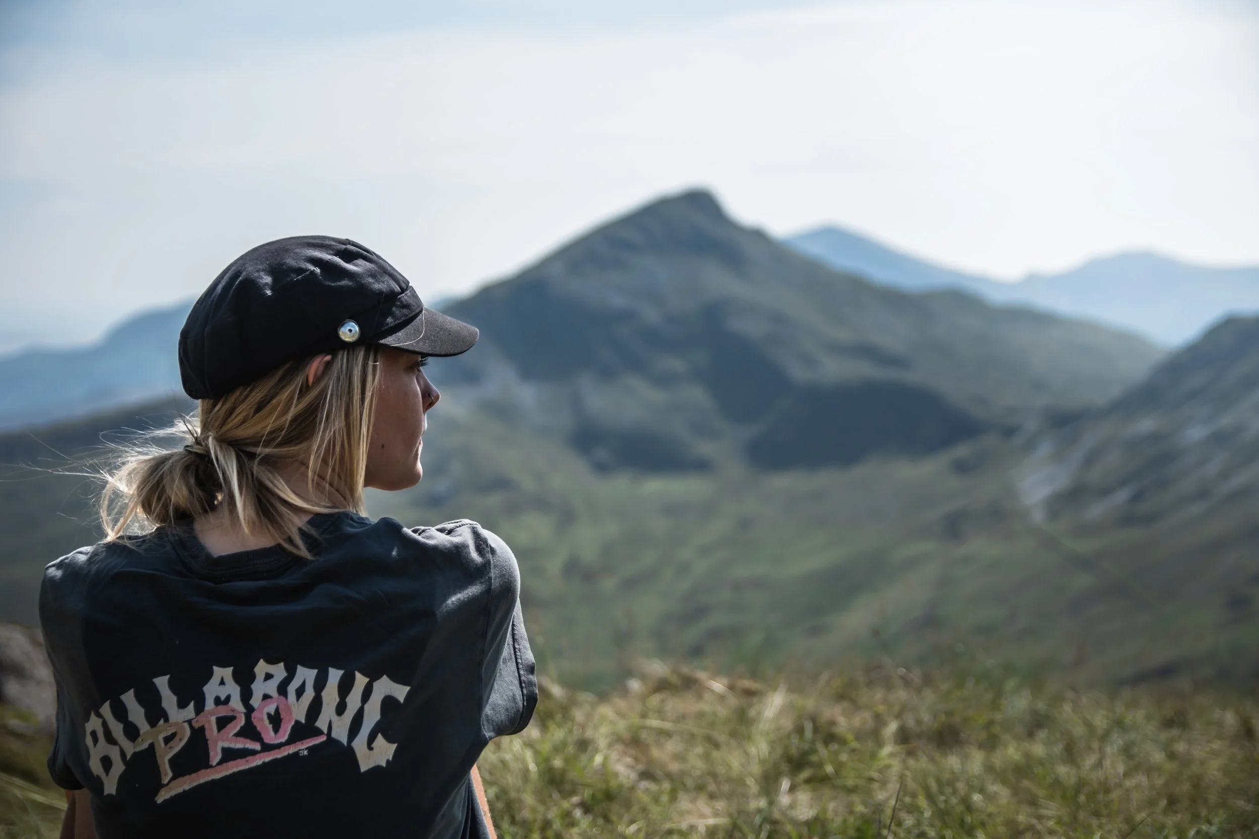 A woman sitting outdoors in front of a mountainous landscape, wearing a black cap and a black jacket with the word 'HILLARIOUS' on the back.